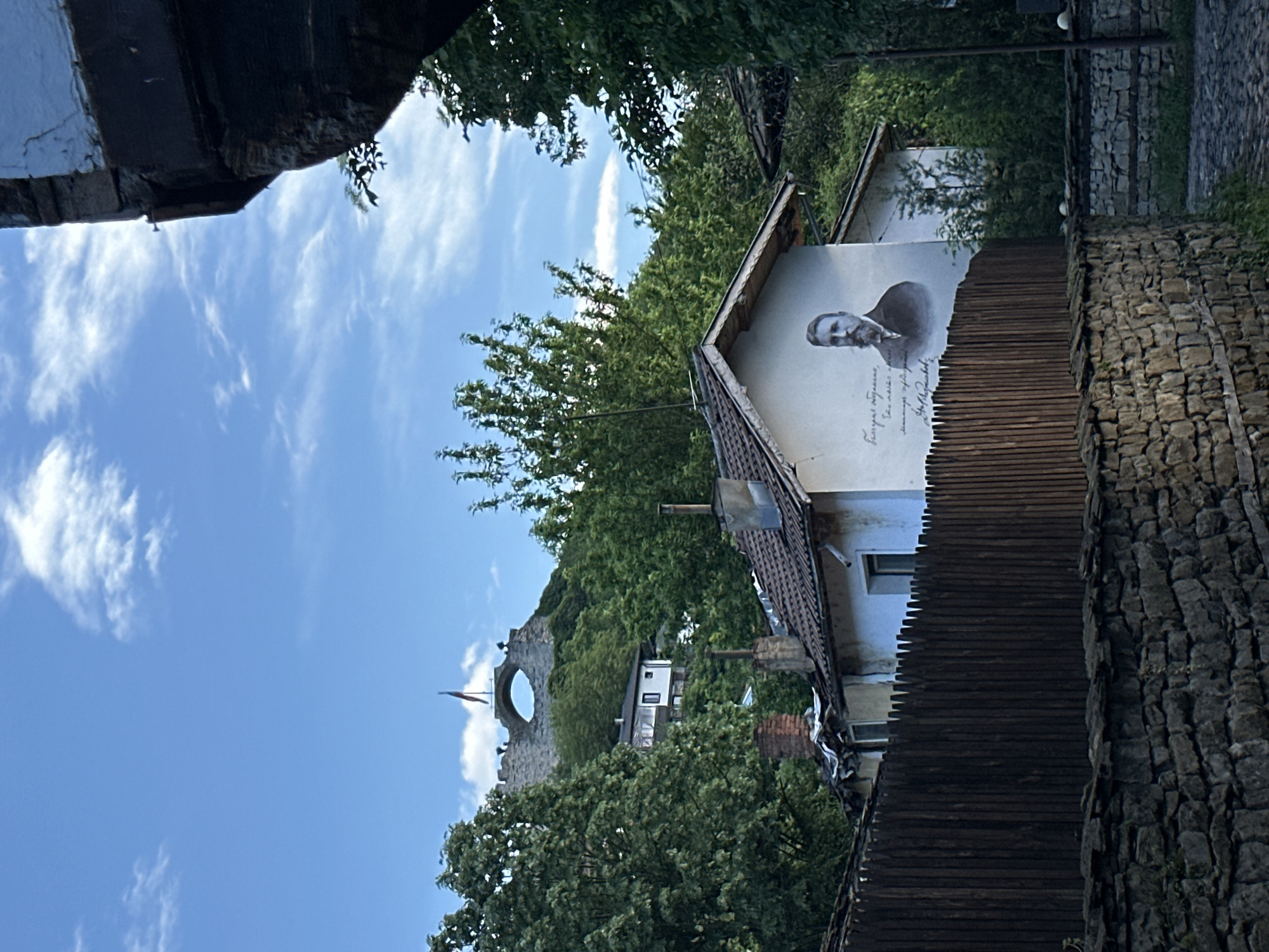 The image shows a street scene in Lovech, Bulgaria, looking towards the medieval Lovech Fortress on a hill. The sky is blue with scattered clouds. A portrait mural of Vasil Levski is painted on the side of a building to the right. A pedestrian walkway constructed from wood is also to the right. Trees are interspersed throughout. A partial view of what appears to be a stone building is in the upper left corner.
