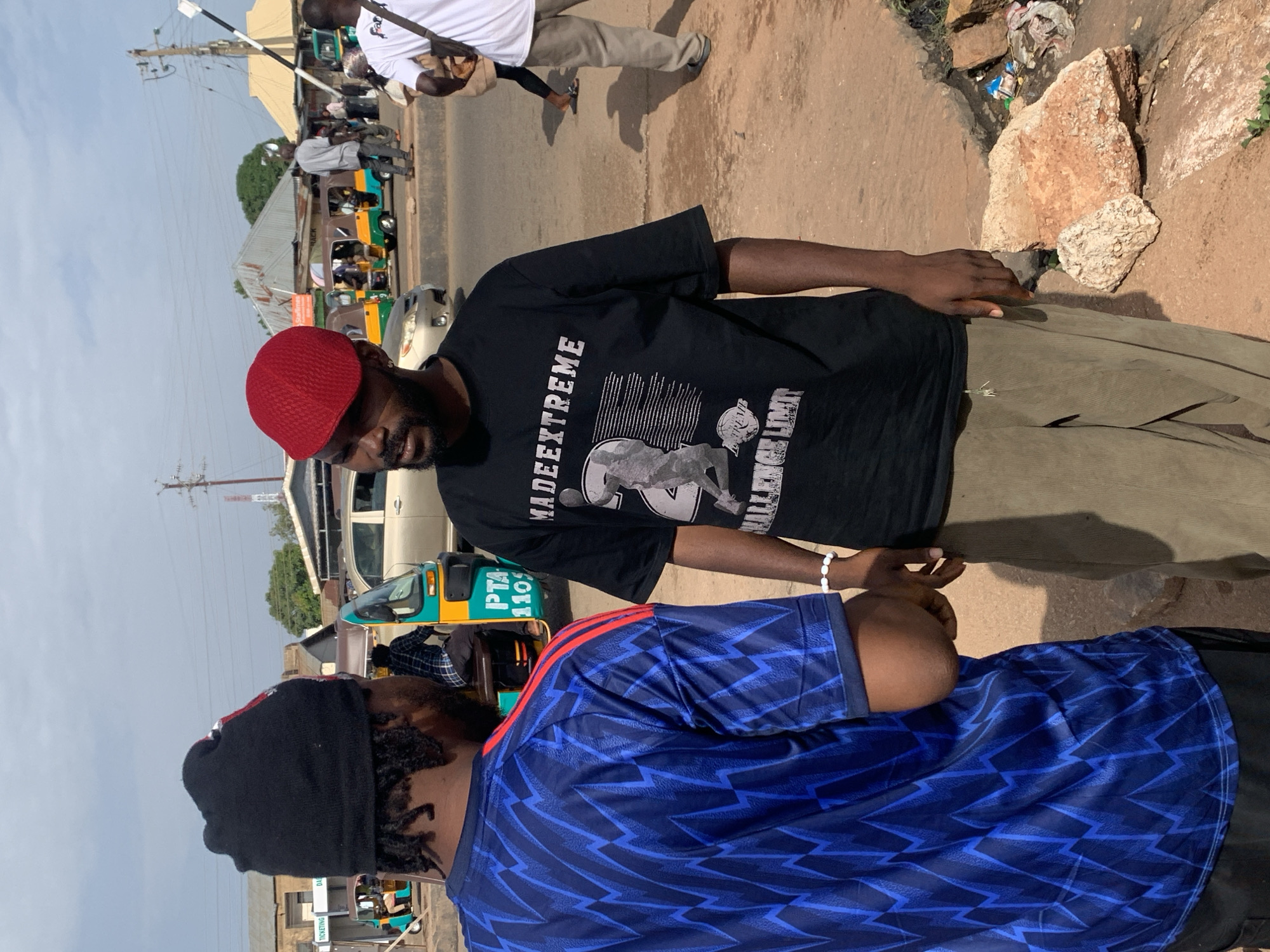 Two men are standing on a dirt road next to each other, facing each other in conversation. The man on the left is wearing a black beanie, a blue shirt with a lightning bolt pattern, and a bracelet. The man on the right is wearing a red beret, a black t-shirt with a graphic of a person and the words "MADEEXTREME" and "CHALLENGE LIMIT", and khaki pants. In the background, there are several auto-rickshaws, buildings, and a power pole. The scene appears to be a busy street in Gura Top, Nigeria, with people and vehicles present. The lighting suggests it is daytime.