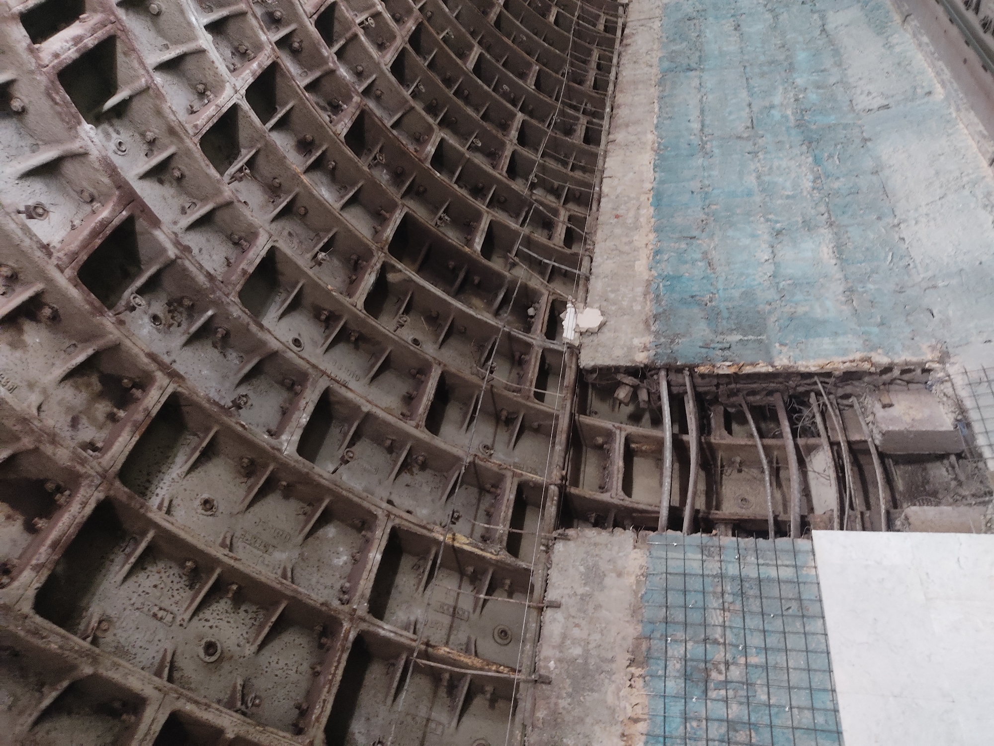 The image shows a section of a tunnel under construction in an unknown location, likely a subway or similar underground infrastructure. The tunnel's wall is partially finished, with a segment composed of interlocking, gray, cast-iron segments bolted together to form a curved, grid-like pattern. To the right, the wall reveals the underlying structure, with steel rods and a wire mesh visible beneath a light blue and gray surface. A rectangular white tile is set against the bottom right corner. The overall impression is that of an ongoing project, revealing the layers of construction of an underground tunnel.