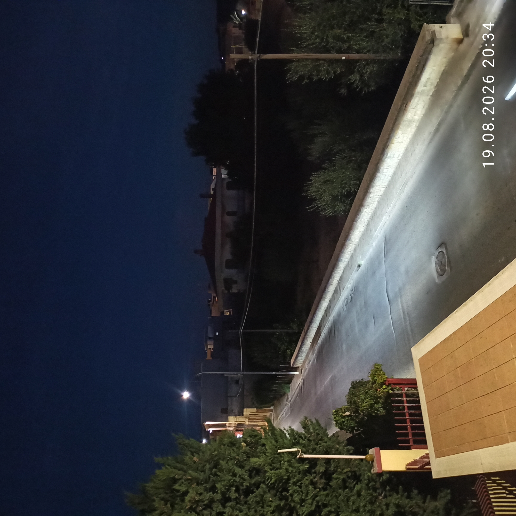 The image depicts a street scene at night in Bitonto, Italy. A street lamp illuminates a paved road that curves to the left. On the left side of the road, there is a tall evergreen tree and a yellow pillar with a red fence. Buildings are visible in the background, some with illuminated windows. On the right side of the road, there are trees and a low stone wall. A manhole cover is visible in the center of the road. The date and time stamp "19.08.2026 20:34" is present in the lower right corner of the image. The overall atmosphere is quiet and still, characteristic of a residential street at night.
