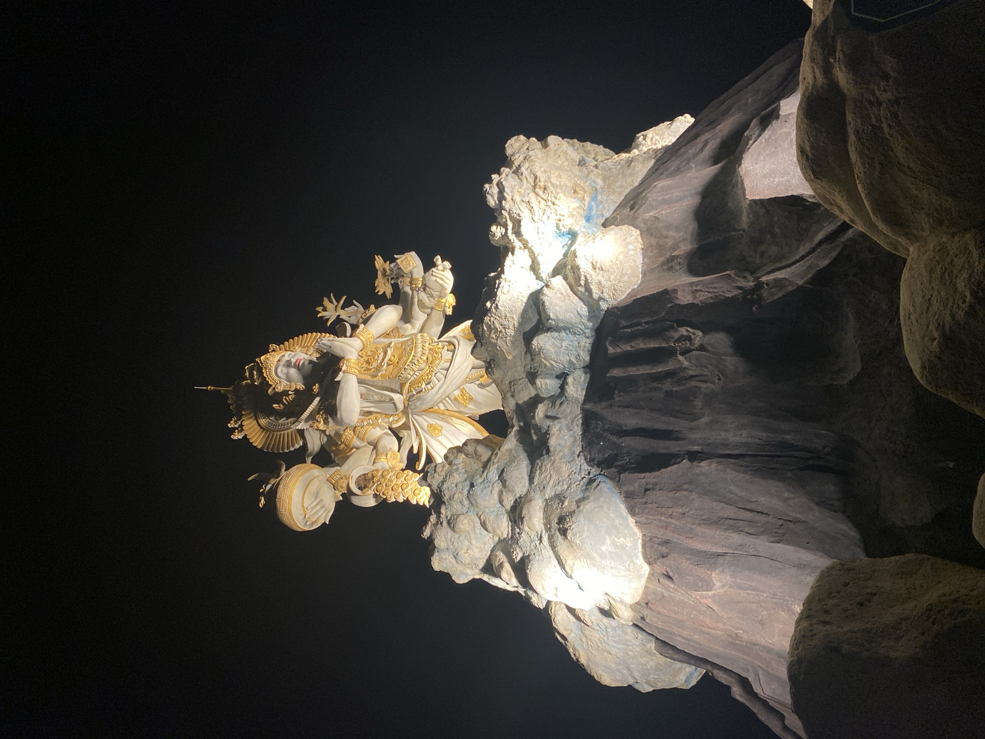 The image shows a statue of a Hindu deity mounted on a rock face, likely within a cave. The statue is predominantly white with gold accents and is intricately detailed. The rock face is multi-toned, ranging from light gray to dark brown, suggesting varying levels of shadow and weathering. The background is solid black, indicating that it is nighttime or a very dark interior space.
Based on the statue's style and the rock formation, this scene is likely from Garuda Wisnu Kencana Cultural Park, located in North Kuta, Indonesia. The statue is one of the depictions found inside.