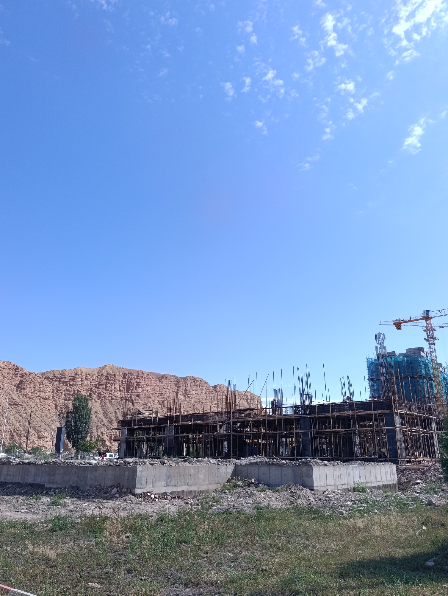 The image shows an under-construction building in Naryn City, Kyrgyzstan. The building is made of concrete and steel rebar, with scaffolding surrounding it. A yellow construction crane is visible to the right of the building. In the background, there are mountains and a clear blue sky with some white clouds. There are several construction workers on the scaffolding. The ground in front of the building is covered with grass and debris.