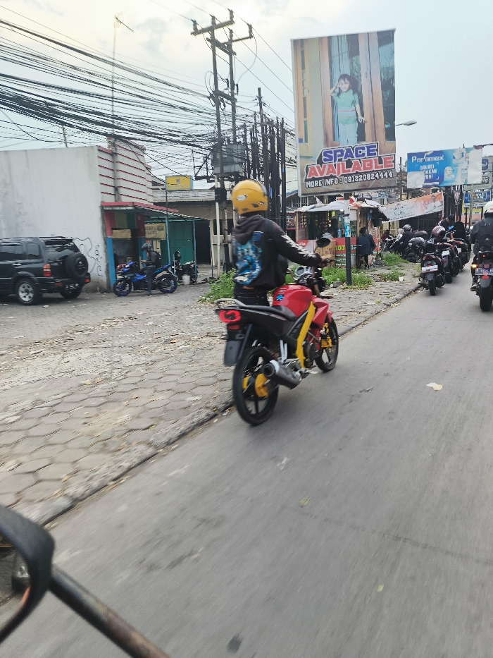 The image was taken from a moving vehicle on a street in Jatinangor, Indonesia. It shows a man wearing a yellow helmet, riding a red and black motorcycle, facing the camera. The road is paved with brick and has a light grey asphalt surface. The man is driving on the right side of the road. A large billboard advertising "Space Available" is on the right. There is a black SUV parked on the left side of the street. Several other motorbikes can be seen in the distance. The road is lined with buildings on both sides, and numerous powerlines are overhead. There is a person standing on the left side of the image, near the SUV. A blue motorcycle is parked in the background. The man riding the red motorcycle has his left arm outstretched and appears to be giving someone a wave or a gesture. It is a bright day and the sky is overcast. The scene is typical of a busy street in an urban environment.