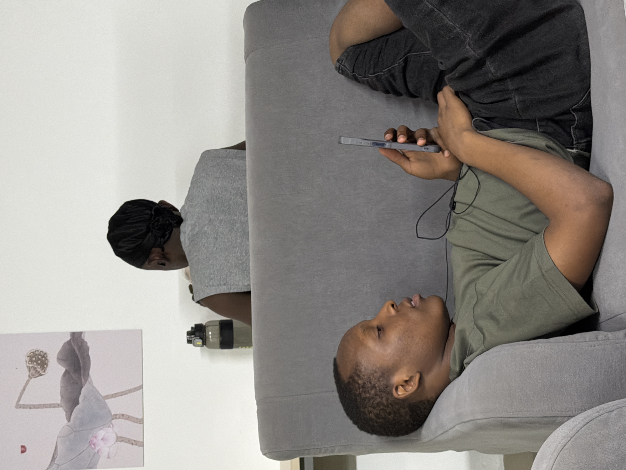 An indoor scene in what is specified as Jiangjin, China, displays two individuals within a domestic environment. In the foreground, positioned on the left, a person with dark skin and short dark hair reclines on their left side on a gray upholstered sofa. They wear a green short-sleeved t-shirt and dark gray shorts. This individual holds a light gray smartphone in their right hand, with black wired earphones connected and draped across their chest; an earbud is visible near their left ear. Their gaze is directed towards the phone screen.
In the background, behind the sofa and partially obscured, a second individual with dark skin is visible from the back. This person is seated at a light-colored surface, possibly a desk, and wears a gray sleeveless top. Their dark hair is styled or covered with a black headwrap. To the right of this individual, on the desk surface, rests a dark translucent water bottle with a black lid and a black band.
On the white wall in the upper left quadrant, a framed piece of art depicting botanical elements, specifically lotus flowers, leaves, and a seed pod, is mounted. The overall environment is illuminated by ambient light, and the walls are plain white. No direct interaction between the two individuals is observable.
