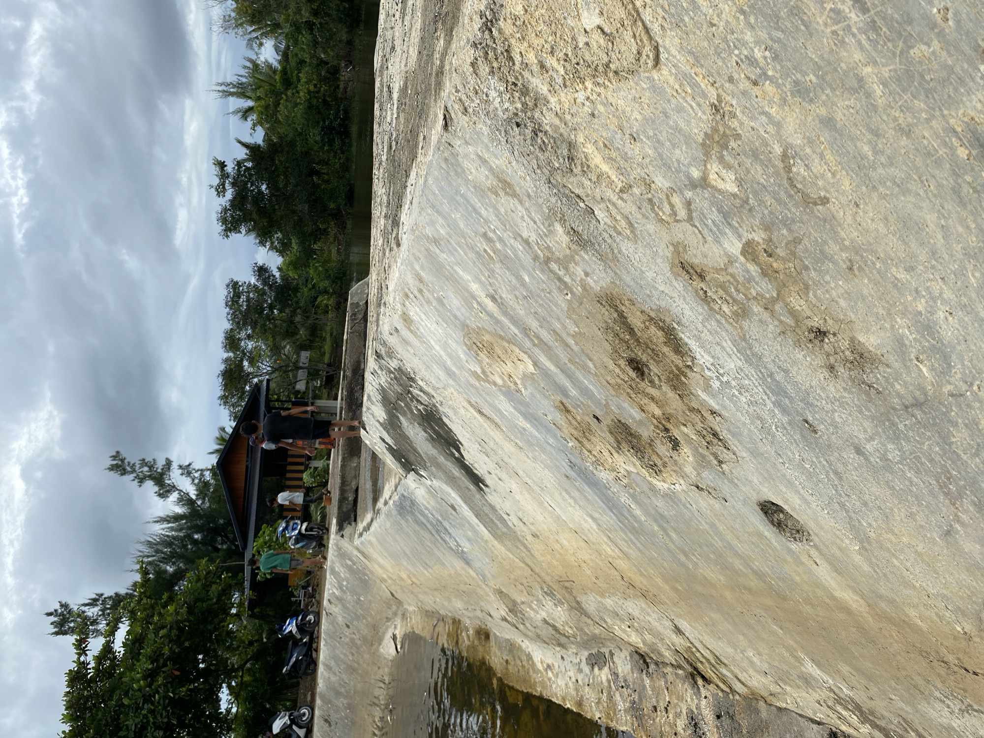 The image depicts a concrete dam structure situated in the city of Aneuk Paya, Indonesia. On the dam's walkway, three young males are visible. Two of them are facing away from the camera, one in a green t-shirt and the other in a white t-shirt. The third individual, wearing a black t-shirt and shorts, is also facing away from the camera but appears to be looking towards the right side of the frame. To the left of the dam, several motorcycles are parked alongside a row of trees. In the background, a rustic wooden structure with a dark roof is nestled among dense green foliage. The sky is overcast with heavy clouds, suggesting a potentially rainy or gloomy day. The dam itself is constructed of rough concrete, showing signs of wear and tear with stains and some discoloration. Water is visible on the left side of the dam, indicating a body of water it is holding back. The overall scene suggests a casual outdoor setting in a rural or semi-rural area.