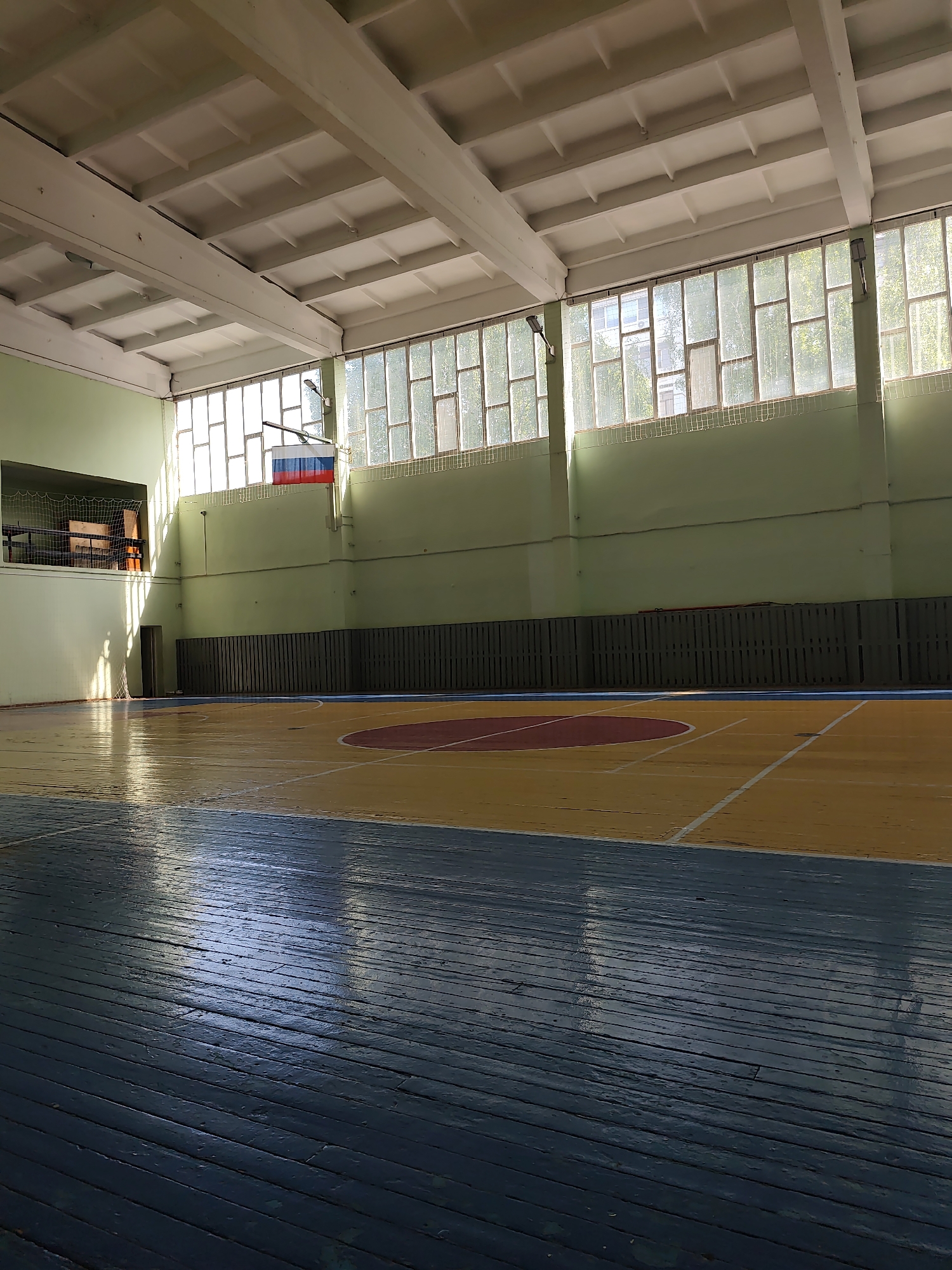The image depicts the interior of a gymnasium in Chaikovsky, Russia. The floor is a hardwood surface with distinct color zones: a blue-painted area in the foreground, transitioning to a yellow-painted area with white basketball court lines, and a central red circle. The walls are painted light green with a horizontal dark wood slat section running along the middle. A gallery with netting is present on the left. Windows run along the upper portion of the far wall, allowing natural light to enter. A Russian flag is affixed to the wall. The ceiling is white and composed of a grid of concrete panels with beams. The room appears empty of people and active participants.