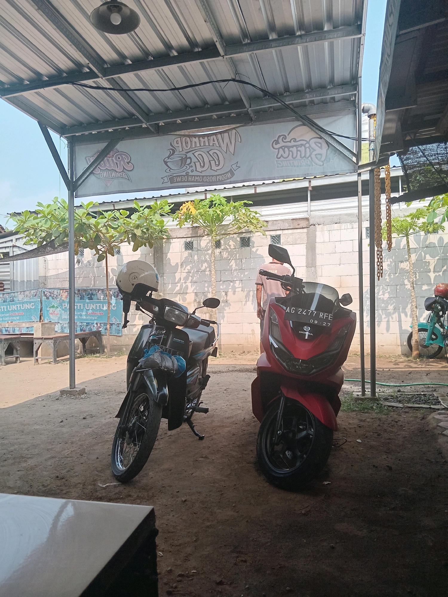 Two motorcycles are parked outdoors under a metal roof. The motorcycle on the left is dark-colored and has a white helmet resting on its handlebars. The motorcycle on the right is red and features a license plate reading "AG 2477 RFE 03-27". A person is standing behind the red motorcycle, facing away from the camera. In the background, there is a white brick wall and some greenery. A banner is hanging from the roof with text that is partially obscured and mirrored. The ground is composed of dirt and gravel. The setting appears to be a roadside eatery or rest stop in Tulungagung, Indonesia, given the common style of outdoor shelters and the visible license plate format.