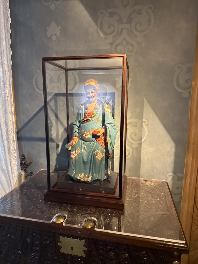 A framed glass display case containing a statue of a seated Asian man wearing a robe is prominently featured. The statue, dressed in teal and gold, is placed atop a dark, ornately carved wooden chest, the surface of which is partially covered with a clear protective layer. The backdrop is a textured, light blue wall decorated with a repeating floral motif. The scene is indoors. The image was captured inside the Pena National Palace in Sintra, Portugal.