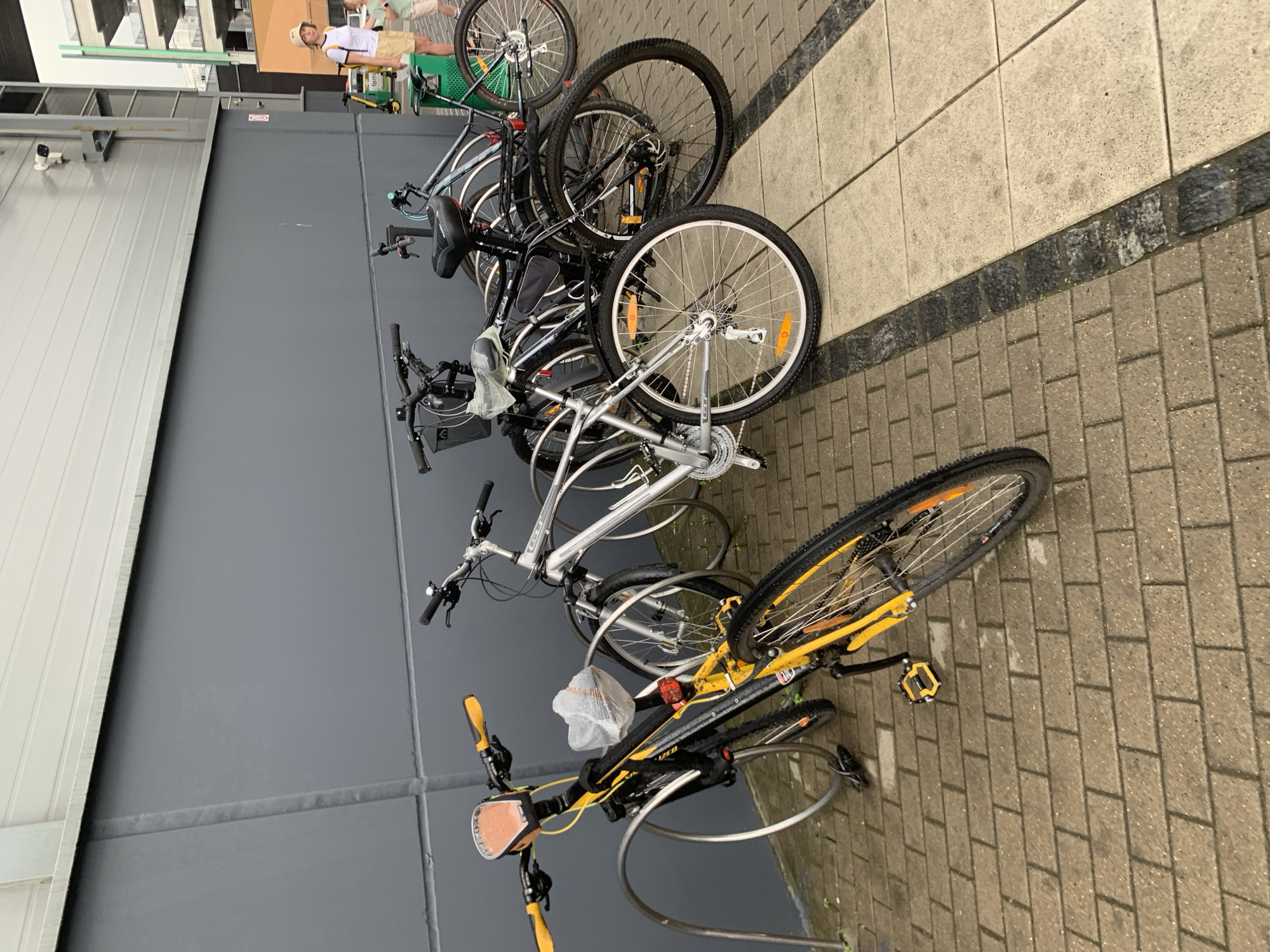 A line of parked bicycles is situated against a dark gray building facade. The bicycles are of various models and colors, with the closest one being a black and yellow mountain bike. To the right of the bicycles, a woman wearing a white t-shirt and a light-colored hat is visible, standing near a green trash bin. She appears to be looking towards the right side of the frame. The ground is paved with brick and stone tiles. In the background, there are parts of other buildings, including balconies and railings. The scene suggests an outdoor urban environment, possibly near a commercial or residential area. The image was captured in Minsk, Belarus.