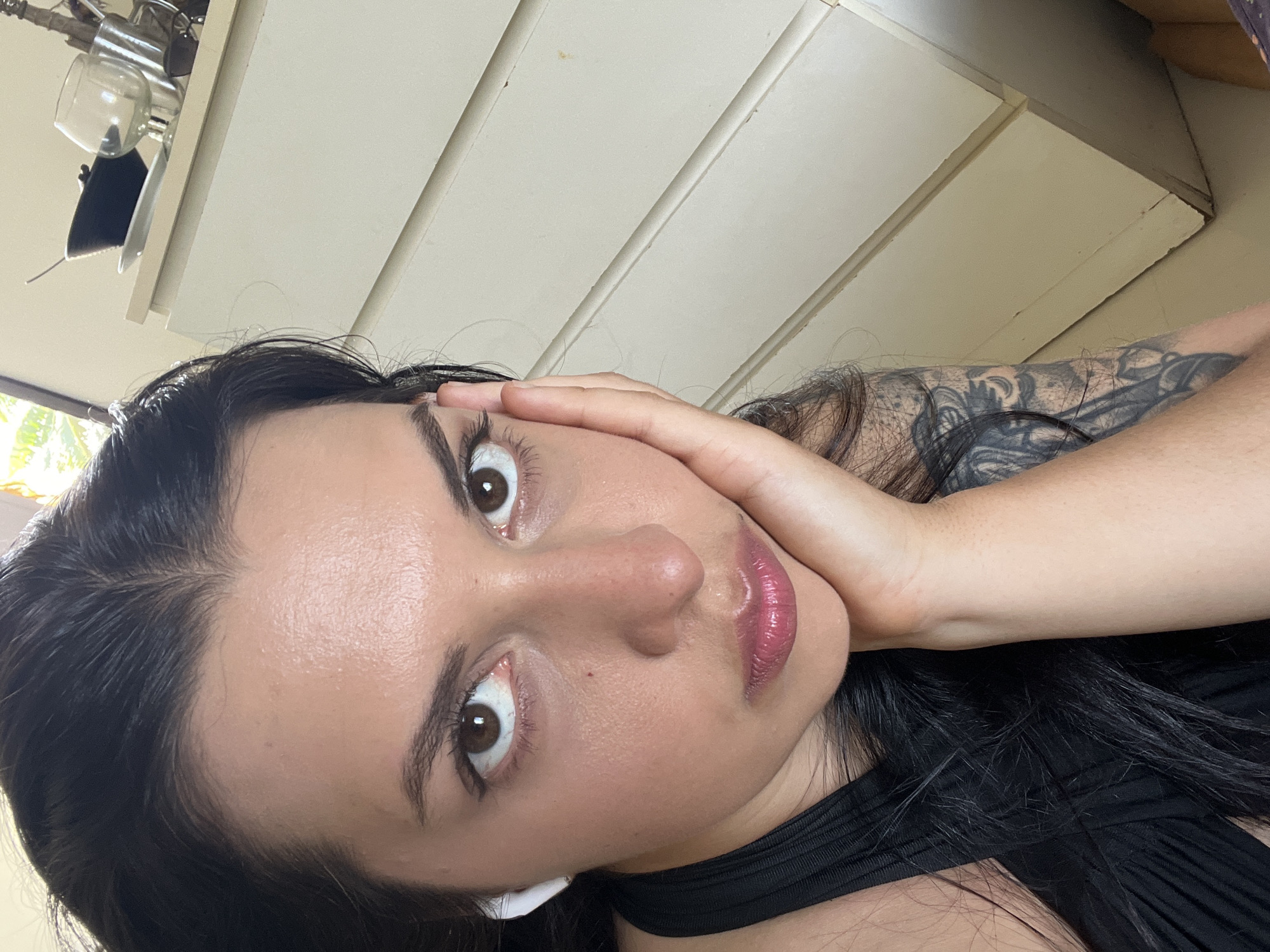 A close-up, head-and-shoulders shot of a woman, taken from a high angle. Her face is tilted and she is looking towards the camera with wide, brown eyes. Her left hand is resting on her cheek, and her fingers are spread out. She has dark, long hair. She is wearing a black top and white earbuds. Her left arm, which is visible in the foreground, has a tattoo. The background consists of a pale, textured wall and what appears to be kitchen cabinetry. The lighting suggests it is daytime. There is no visible text in the image.