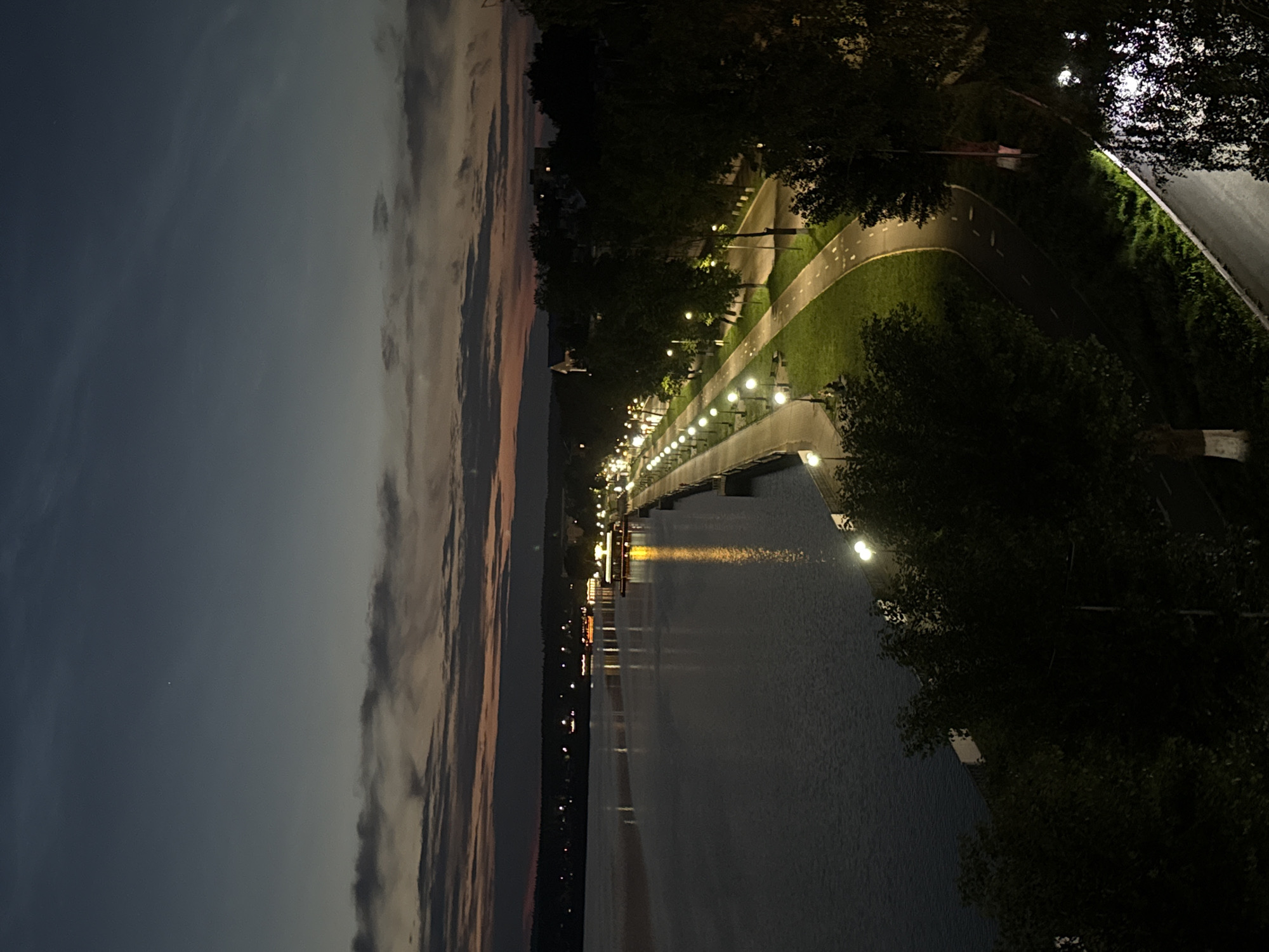 The image depicts a nighttime scene of a waterfront promenade in Votkinsk, Russia. The sky is a deep blue, transitioning to orange and pink hues near the horizon, suggesting either dusk or dawn. Wispy clouds are visible. A large body of water, likely a lake or river, occupies the left side of the frame, reflecting the lights from the opposite shore.
Along the waterfront, a paved promenade is illuminated by numerous lampposts, casting a warm glow. A walking path and a separate bike path run parallel to the water's edge, bordered by a strip of green grass and trees. Buildings and lights are visible in the distance across the water, indicating a populated area. The right side of the image shows trees and a street or road illuminated by streetlights. No people are clearly visible in the image, and no specific actions or interactions are depicted. The overall atmosphere is serene and tranquil, with the artificial lights creating a striking contrast against the natural darkness of the night.