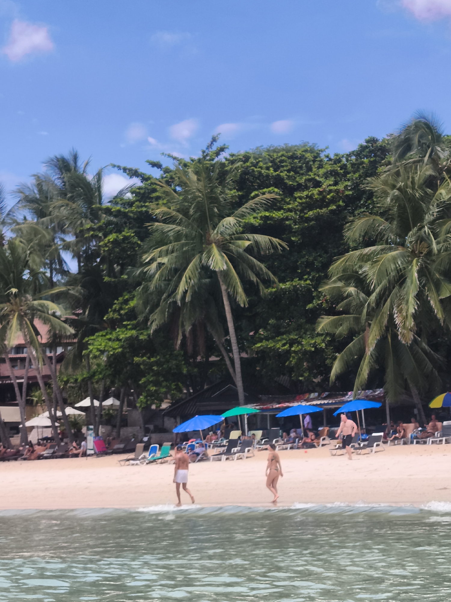 The image depicts a beach scene in the Ko Samui District, Thailand, under a bright blue sky with scattered white clouds. Palm trees with dense green foliage line the background, with some extending over the sandy beach. The beach is populated with numerous beach chairs, some occupied by sunbathers, and several colorful umbrellas providing shade. In the foreground, two people are walking near the waterline. A man, wearing white shorts, is walking away from the viewer towards the right. A woman, wearing a bikini, is walking towards the viewer from the right. The ocean in the foreground is calm with gentle waves lapping the shore. Buildings and more vegetation are visible in the background beyond the beach chairs and umbrellas.