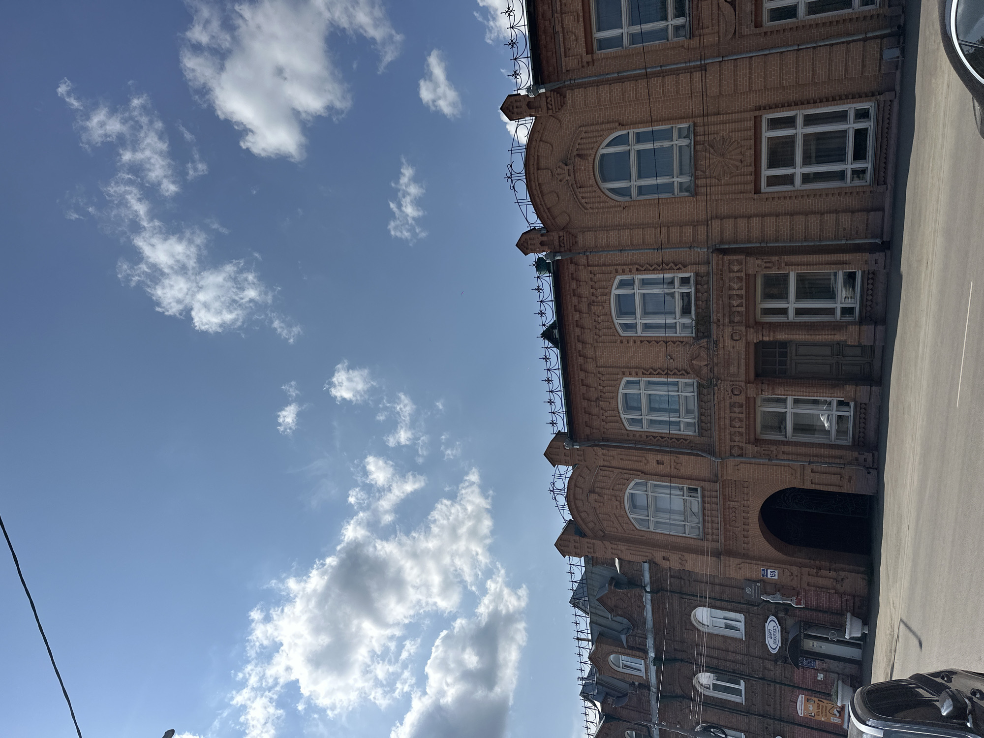 The image depicts a street scene in Biysk, Russia, on a sunny day with a blue sky and scattered clouds. The foreground shows the asphalt road and the front of several brick buildings, some with arched windows and decorative facades. One building features a large arched entrance. Signs in Russian are visible on some of the buildings. The architecture suggests an older style. A black car is partially visible on the left side of the image. The overall impression is that of a historical urban setting.