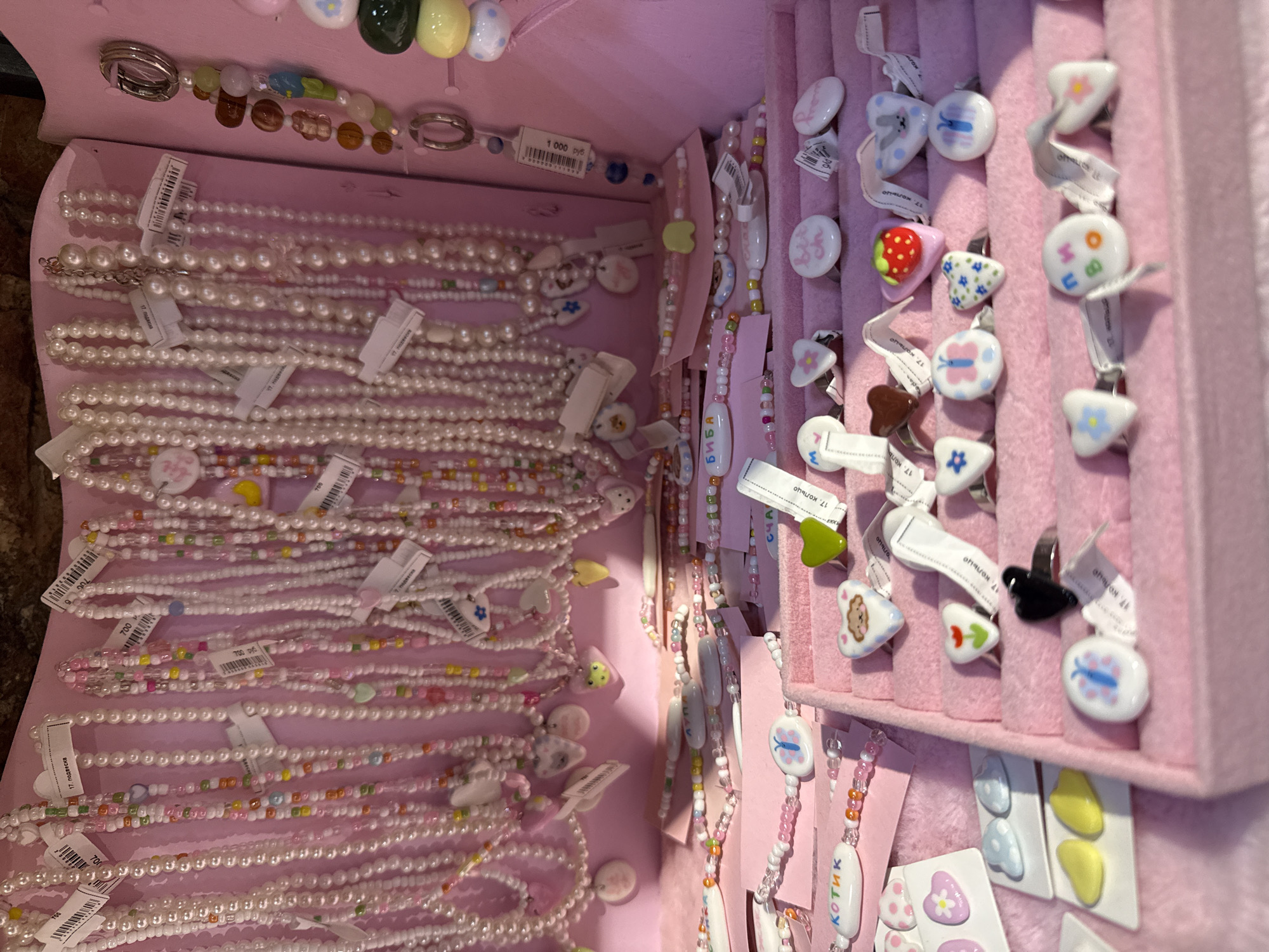 The image shows a display of colorful jewelry, primarily necklaces and rings, inside a pink jewelry box. The items are predominantly made of beads, pearls, and ceramic charms. Some of the charms are heart-shaped, while others feature animals, fruits, or flowers. Many of the necklaces have small, colorful beads interspersed with white pearls. The rings are displayed in rows within the velvet-lined compartments of the box. There are also some loose charms and smaller pieces of jewelry on display. The overall aesthetic is playful and childlike, with a dominant pink color scheme. The context of this image being located in Saint Petersburg, Russia is not evident from the visual information alone.