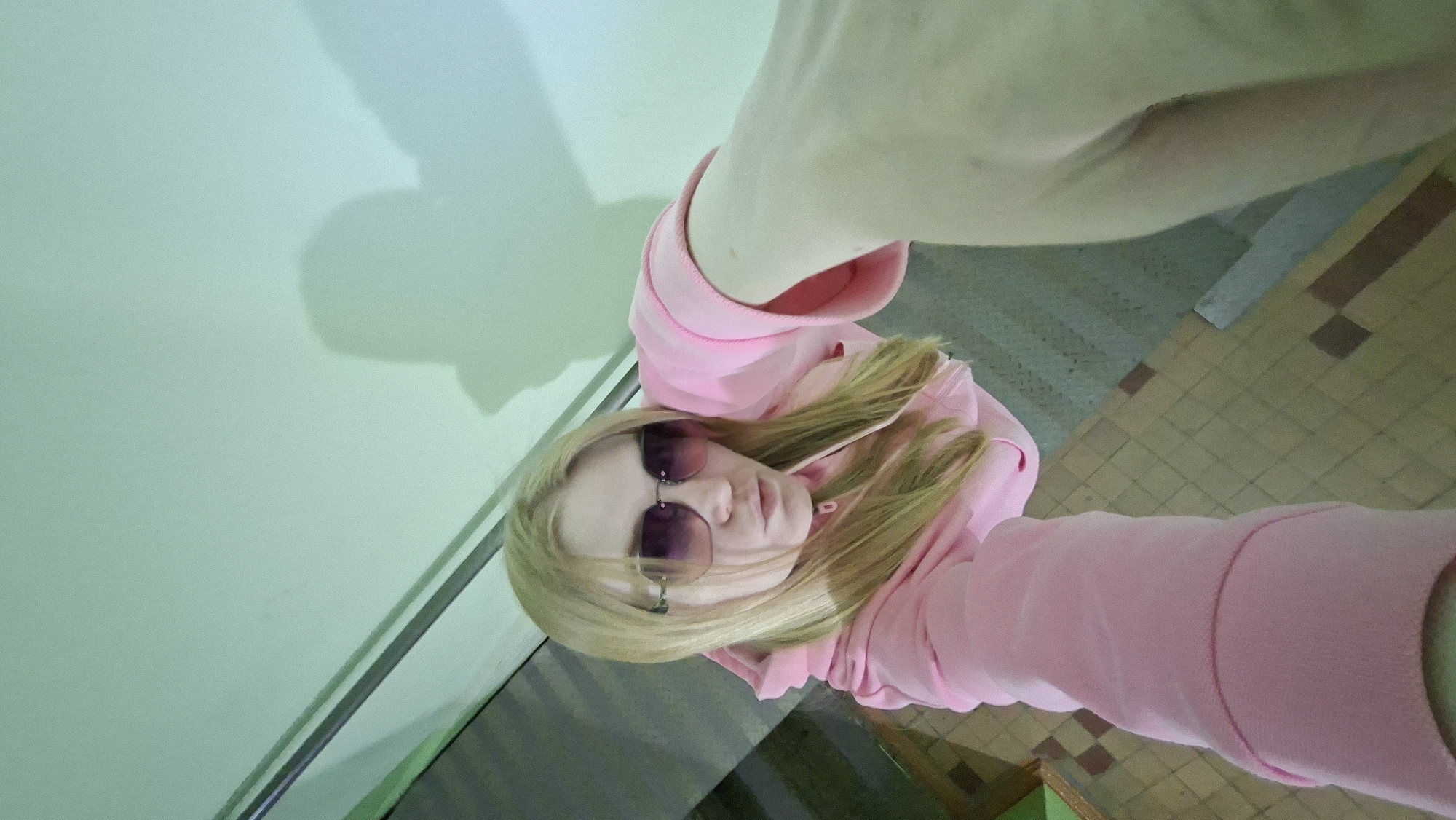 A selfie taken in a room with mint green walls and a tiled floor in Chaikovsky, Russia. A woman with blonde hair is wearing pink sunglasses and a pink hooded sweatshirt. Her arm is extended, holding the camera. The lighting casts a shadow on the wall.
