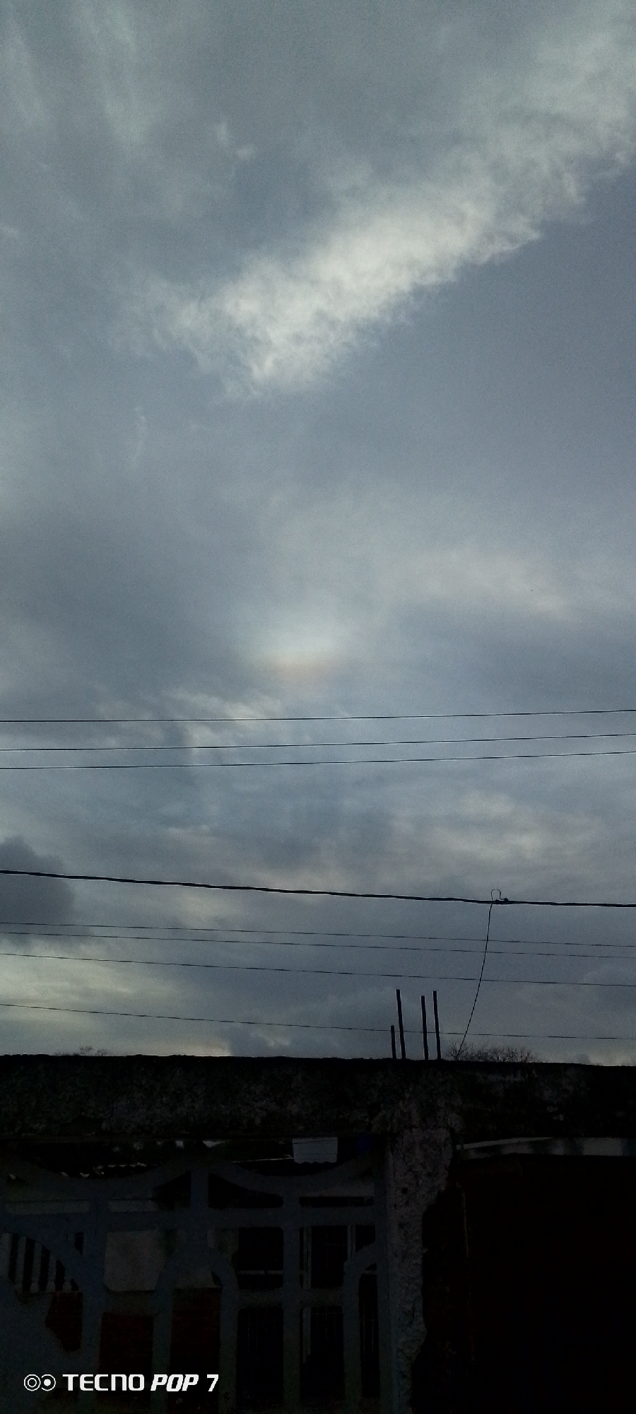 The image shows a gray, overcast sky with a visible sun halo, with power lines running horizontally across the middle ground. Below the power lines is a low building with a partially visible gate and what appear to be thin metal poles extending from the roof. The image was taken in Altagracia, Venezuela. There are no people visible.