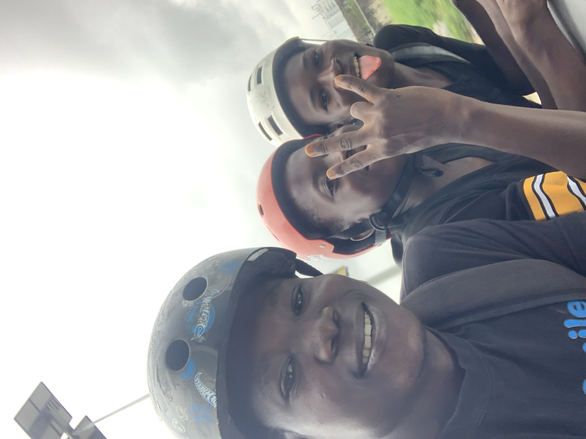 Three young Black individuals wearing helmets are posing for a selfie on a cloudy day in Ikoyi, Nigeria. The person in the foreground, on the left, wears a black helmet with a "Hot Wheels" design and smiles at the camera, revealing their teeth. They are wearing a black t-shirt with the word "Smile" visible in blue lettering. To their right, a person in an orange helmet looks towards the camera and holds up two fingers in a peace sign. Further to the right, another person in a white helmet sticks their tongue out at the camera, also displaying a peace sign with their right hand. They are all looking at the camera. In the background, a lamppost with a solar panel is visible on the left, and a building can be seen in the distance on the right. The overall environment appears to be an outdoor urban setting.