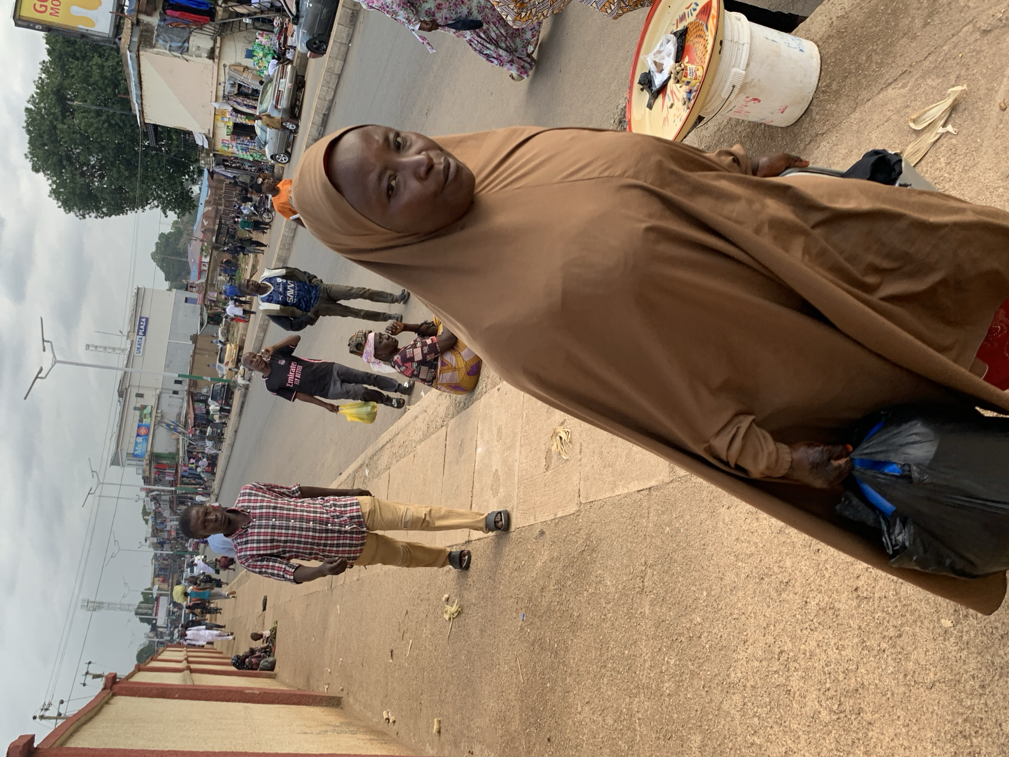 The media file shows a street scene in Jos, Nigeria. In the foreground, a woman in a brown hijab is looking towards the camera, with her arm resting on a brown cloth that drapes over her. Beside her, to the left, a man in a checkered shirt and tan pants is also looking towards the camera, with his hands by his sides. Behind them, a bustling street is visible with several people walking and standing. Shops line the street, and a few vehicles are present.
The scene appears to be during the day, with overcast sky conditions. The overall atmosphere is that of a busy marketplace or commercial area. The clothing of the people suggests a warm climate. The ground is paved with concrete and a rougher textured sidewalk.
In the background, the image shows a longer stretch of street with more shops and people. A sign is visible on one of the buildings, but the text is not clearly legible. Another sign near the top left of the image contains the partial text "GO MO". A large white bucket with a red rim and a tray on top containing some items is placed near the woman in the foreground. A black plastic bag is also visible. The image is shot at an angle, with the foreground subjects appearing to be positioned closer to the camera.