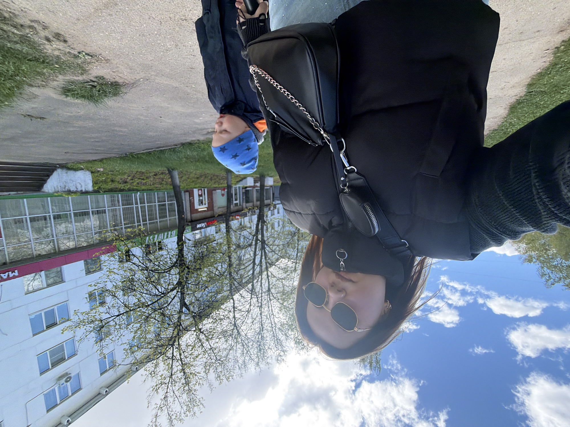 The image shows a woman wearing sunglasses and a black jacket with a black purse, standing next to a young boy wearing a blue hat. They are outside in Chaikovsky, Russia, on a sidewalk next to a building with a red sign that includes the Russian word "МАГ". There are trees in the background and a cloudy sky above. The photograph is oriented at an angle.