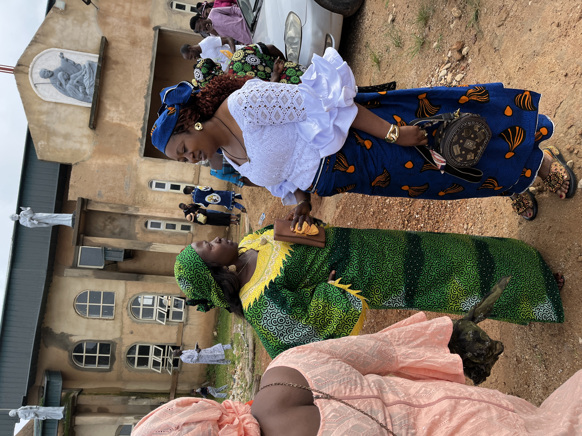 Two women are conversing outside a building that appears to be a church, possibly located in Jos, Nigeria. The woman on the left is wearing a pink dress and the woman on the right is wearing a green and yellow dress with a matching headscarf. Both women are looking at each other and appear to be engaged in conversation. The building behind them is made of light-colored stone and has several windows and statues on its facade. In the background, other people can be seen milling about. The ground is covered in dirt and gravel. The overall scene suggests a social gathering or event taking place outdoors.