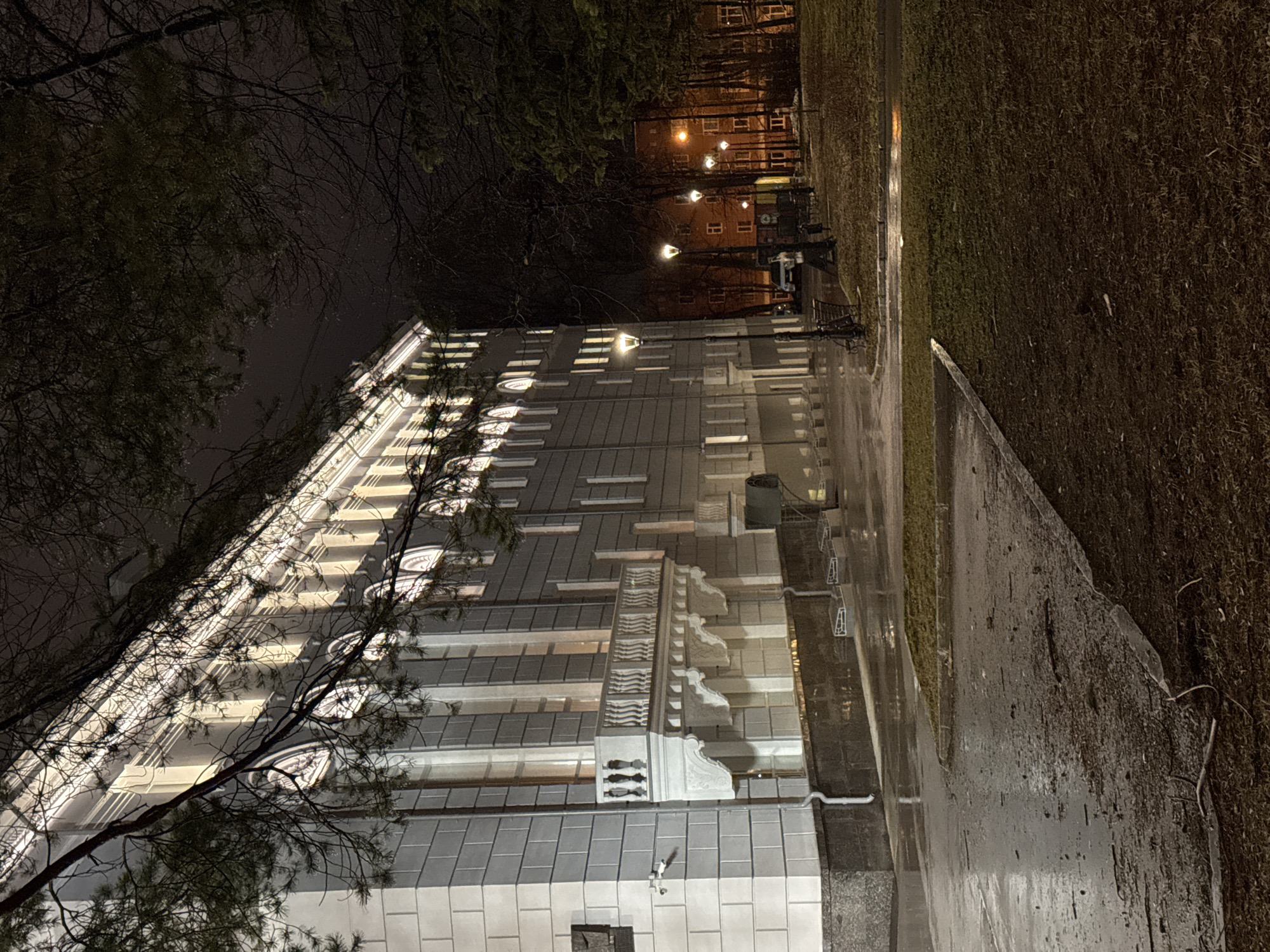 At night, a low-angle view captures a multi-story building with illuminated windows. The building's facade is largely white or light-colored, with distinct horizontal sections. To the left of the frame, tree branches obstruct part of the view, adding depth. A sidewalk or paved area borders the building. Across the sidewalk, there's a grassy area or lawn that transitions into what appears to be a dark, forested area in the distance. Street lamps cast a warm glow, highlighting buildings further in the background. The image might be taken in Perm, Russia, near a large, stately building during an evening.