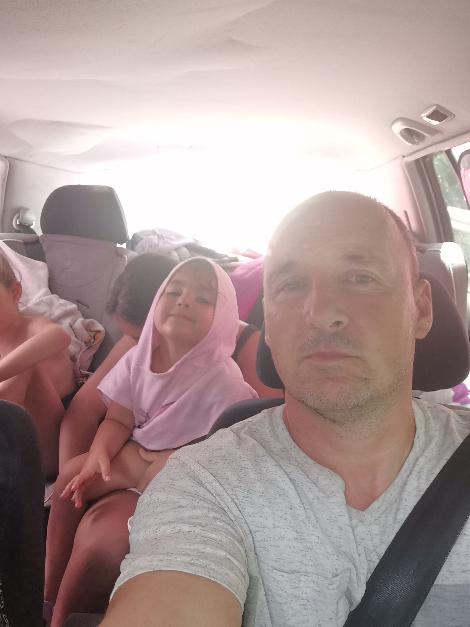 The image shows a man in the front seat of a car, taking a selfie. Behind him, three children are visible in the back seat. The man is wearing a light gray t-shirt with a textured pattern and a black seatbelt is across his chest. He has short, dark hair and stubble on his face. His expression appears neutral or slightly tired.
The children are positioned closely together. The child closest to the camera is a young girl wearing a light pink hooded shirt. She is looking towards the camera and smiling slightly. To her left, another child, partially obscured, is also present. A third child, on the far left, is also visible. The back of the car is packed with luggage, including blankets and what appear to be bags.
The scene is set inside a car, likely during a road trip. The bright light filtering in from outside suggests it is daytime, possibly late afternoon. The interior of the car is somewhat cluttered, indicating travel. The overall mood seems to be one of a family on a journey, with the man capturing a moment of their trip. There is no visible text in the image.