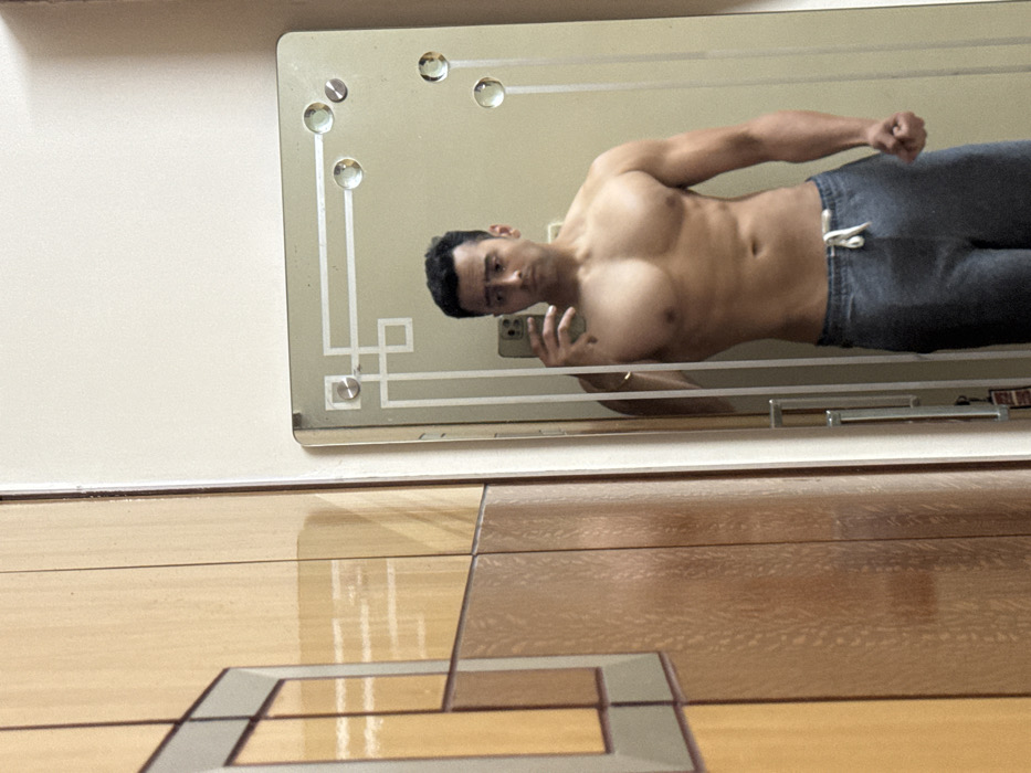 A muscular man with dark hair and gray sweatpants poses for a mirror selfie. He is bare-chested, showing his toned physique. The mirror has an ornate design with geometric patterns and decorative circles. The background features a wall and a floor with a patterned, wooden-tile design. The camera angle is low, emphasizing the floor's perspective. The overall lighting is bright, highlighting the textures of the surfaces and the man's physique.