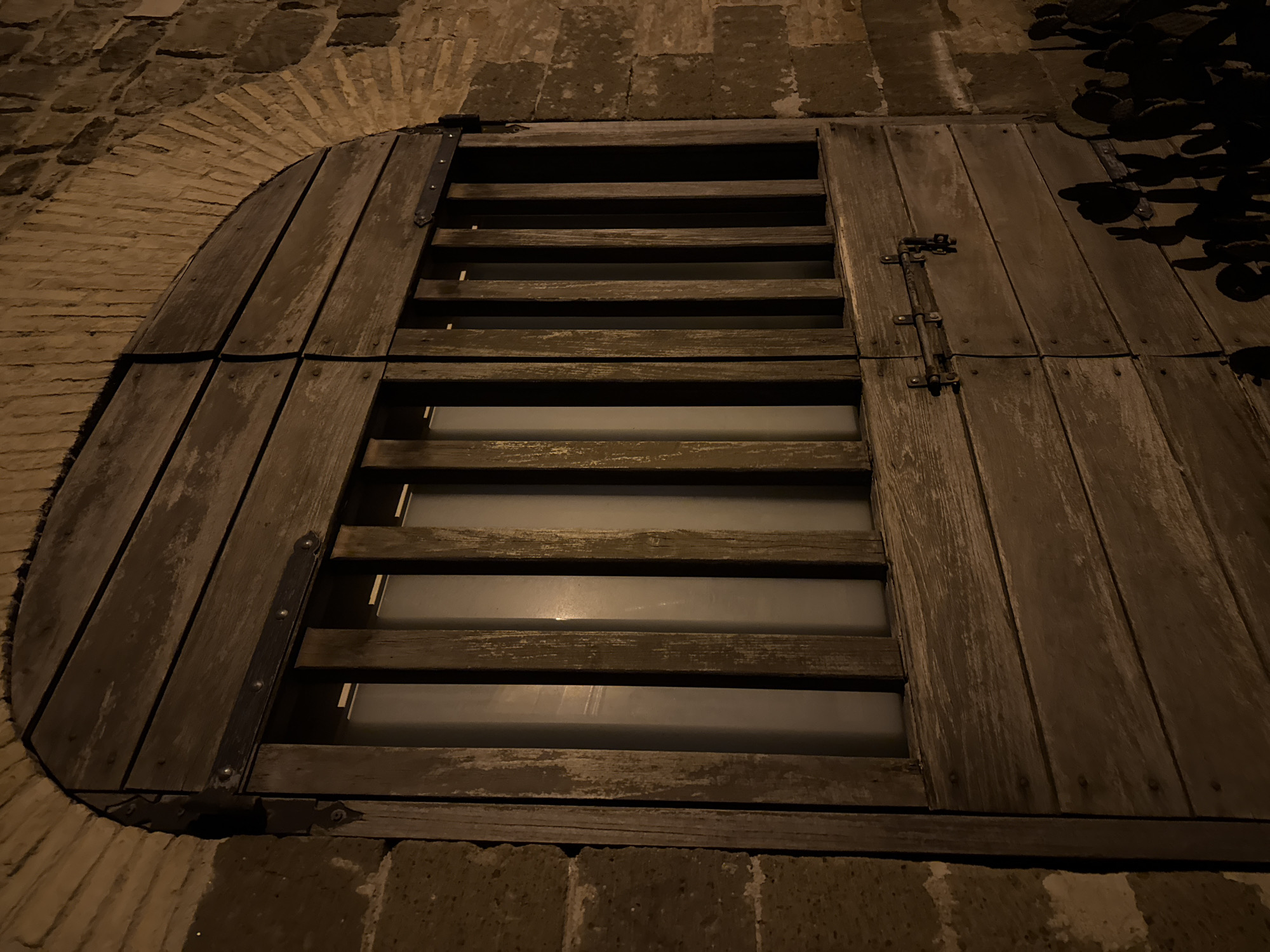The image depicts a wooden, arched window or hatch, possibly a cellar entrance, set into a brick wall. The structure is composed of horizontal wooden slats with gaps allowing light to pass through. A vertical wooden panel runs alongside the slats on the right side. Metal hinges and a latch are visible on the wood. The brickwork surrounding the hatch forms a rounded arch on one side and is darker and rougher on the other. The lighting is dim, with shadows obscuring some details. The location is Castelnuovo di Porto, Italy.