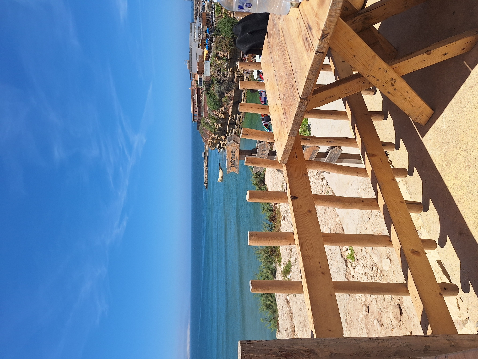 This image captures a scenic view from a wooden picnic table on a rocky terrace overlooking the ocean in Imsouane, Morocco. The foreground features a rustic wooden picnic table and a wooden railing. Beyond the railing, a tranquil blue ocean stretches to the horizon under a clear blue sky with scattered wispy clouds. Along the coastline, rocky cliffs rise, topped with a cluster of buildings, some with red roofs, suggesting a small town or resort area. A small boat is visible in the bay. Several wooden signs are planted in the ground, one pointing to "IMI BAY" and another to "HOTEL". The environment appears to be a popular spot for tourists or locals to enjoy the seaside.