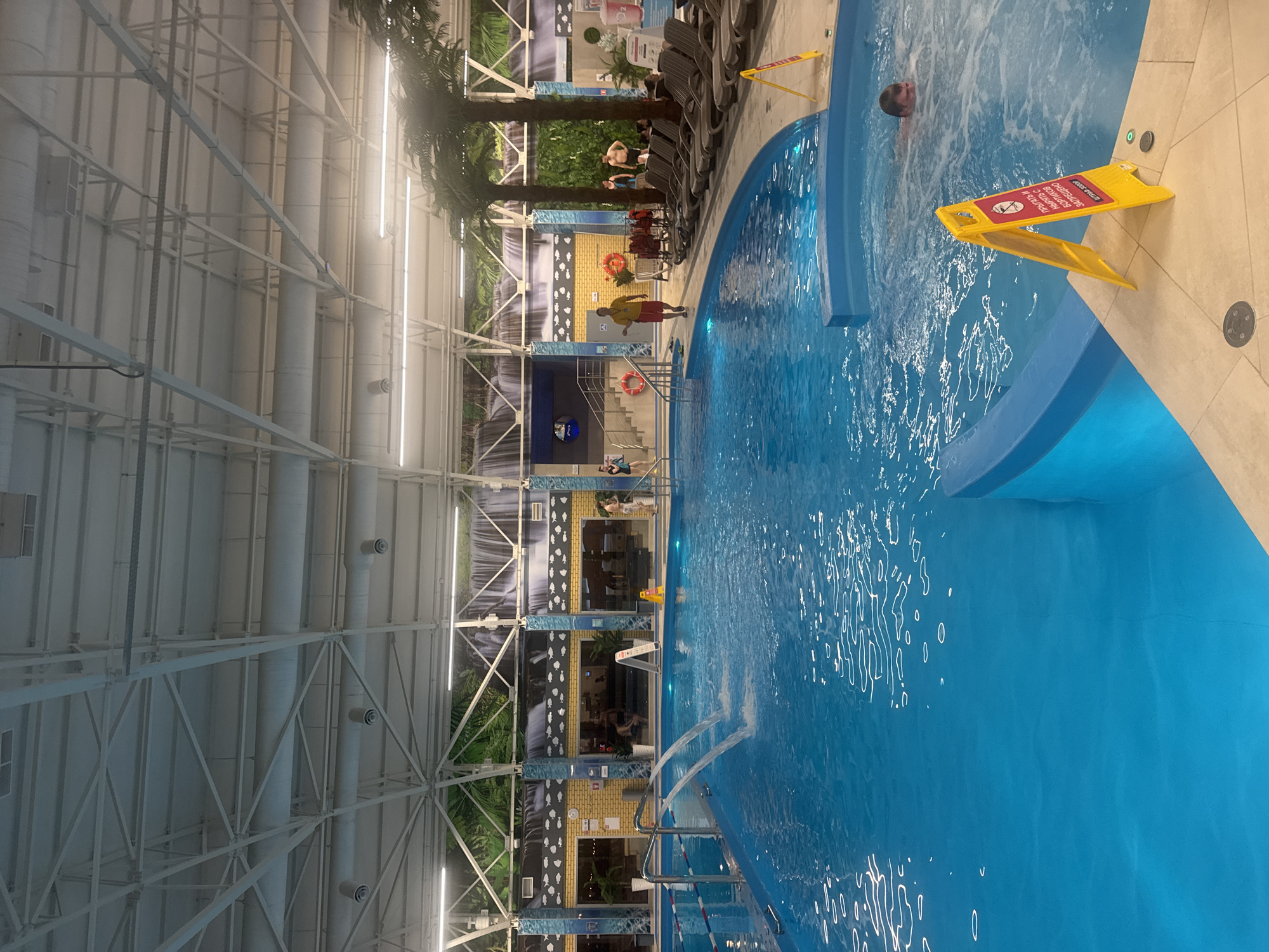 The image shows a water park, likely in Ulyanovsk, Russia, based on the presence of a Russian-language warning sign. A large swimming pool occupies the right side of the frame, with water features including jets or waterfalls. People are swimming in the pool. A lifeguard is positioned near a stairway into the pool, wearing a red and yellow uniform. Numerous deck chairs are arranged near the pool, along with artificial palm trees. The building has a high ceiling supported by a white metal structure, and a mural depicting a waterfall is visible on the back wall.