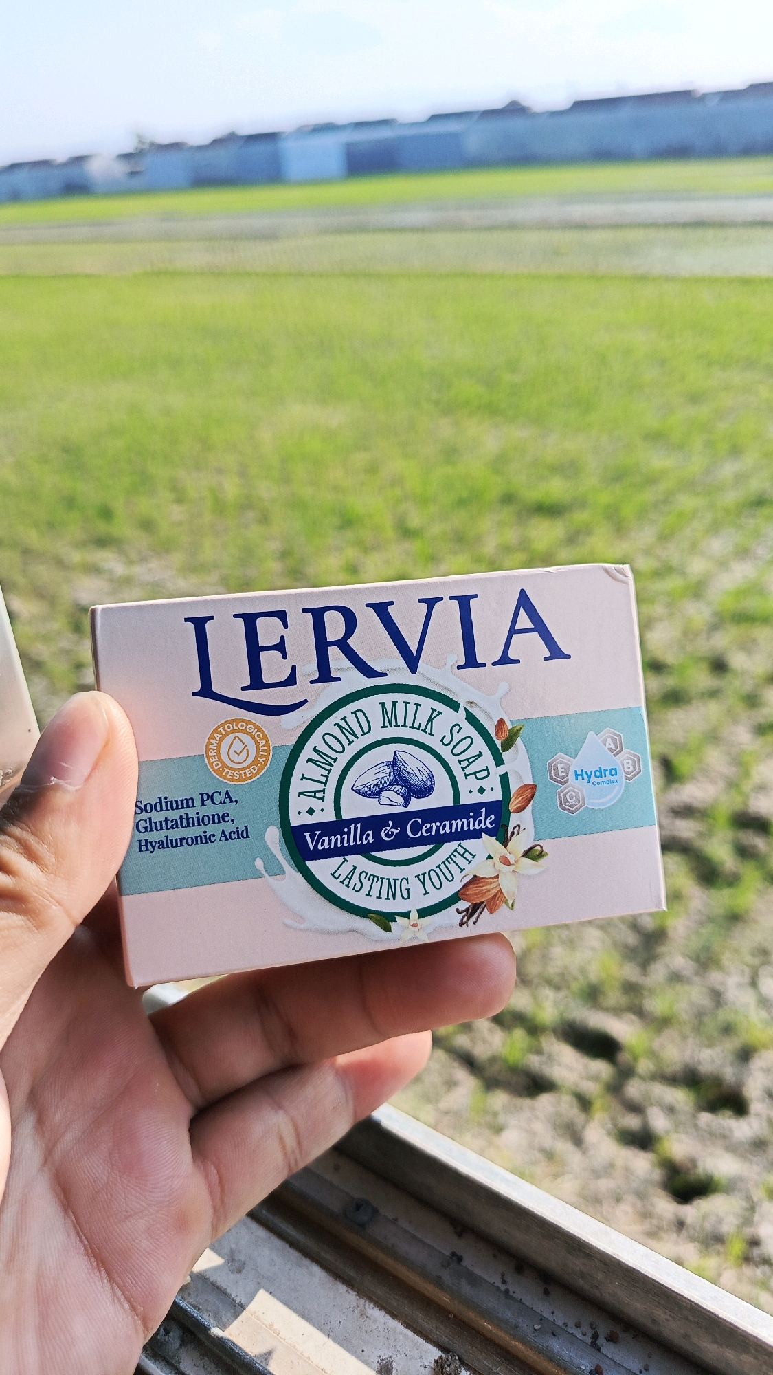 A hand holds a pink and blue soap box in front of a blurred green field with a building in the background. The soap box has "LERVIA" printed in large blue letters at the top. Below this, a circular seal reads "DERMATOLOGICALLY TESTED" and features a stylized almond. The center of the box showcases "ALMOND MILK SOAP" in a circle surrounding an illustration of almonds. Further text states "Vanilla & Ceramide LASTING YOUTH". A small blue box with "Hydra Complex" is also visible. The hand is positioned on the left side of the frame, obscuring part of the soap box. The environment appears to be outdoors with natural light. The location is identified as Babadan, Indonesia.