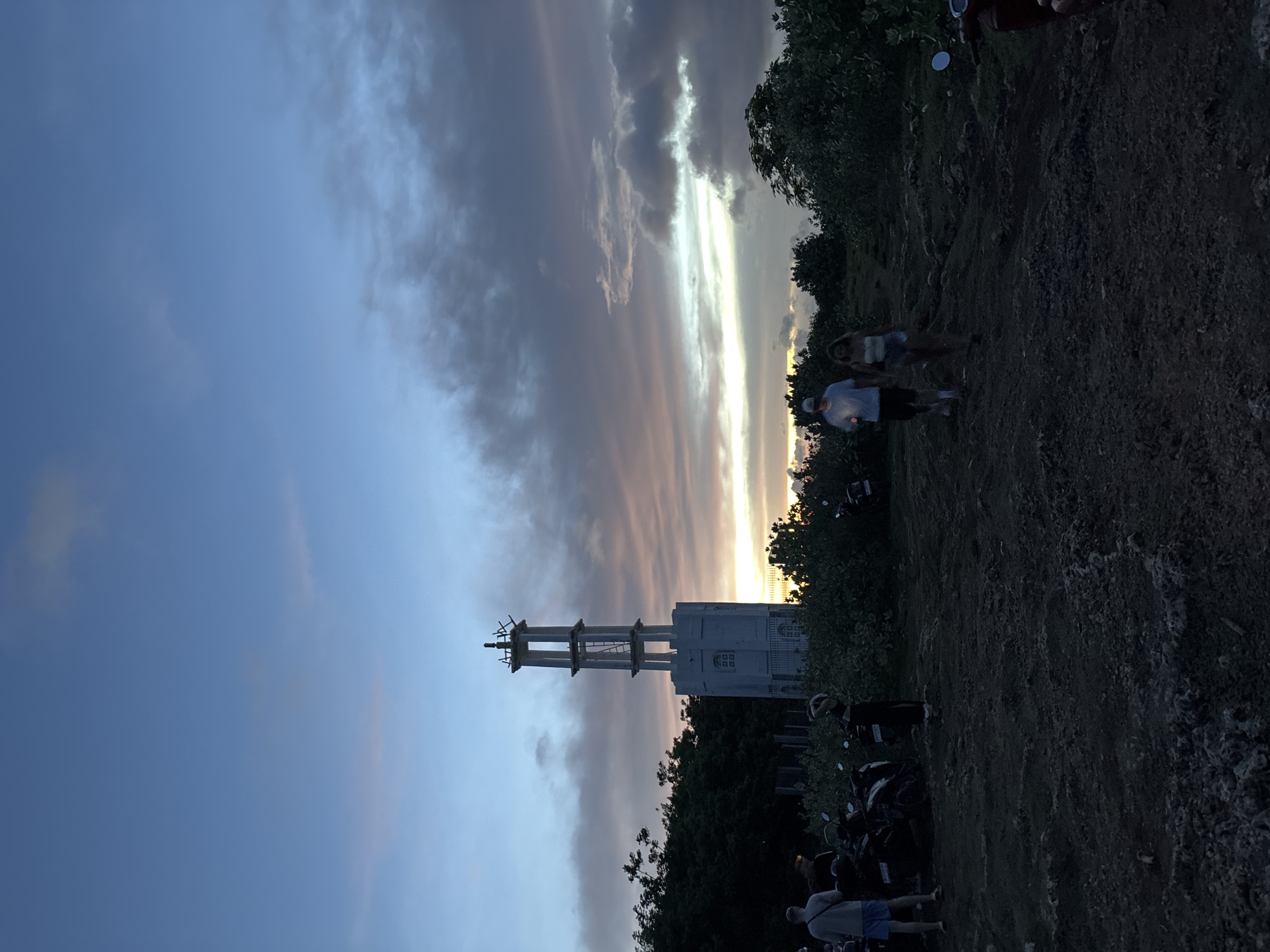 The image captures the Uluwatu Lighthouse in Uluwatu, Indonesia. The lighthouse, a white structure, is positioned in the middle ground, with a partially cloudy sky and a sunset glow behind it. Trees and vegetation surround the lighthouse. In the foreground, there are people standing or walking near parked motorbikes. The ground appears to be rocky.