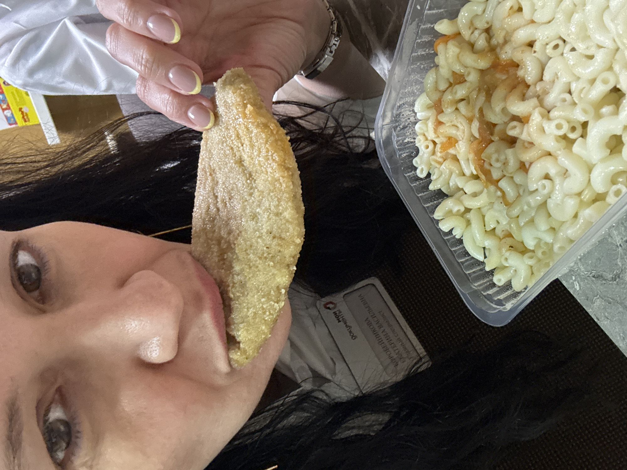 A person with dark hair and light skin holds a breaded food item near their mouth while posing for a photo. A plastic container of macaroni and cheese is visible beside them. The location is likely in Chaikovsky, Russia, as a document card with Cyrillic text and the word "Чайковский" is partially visible in the background. The person has manicured fingernails with a yellow accent.