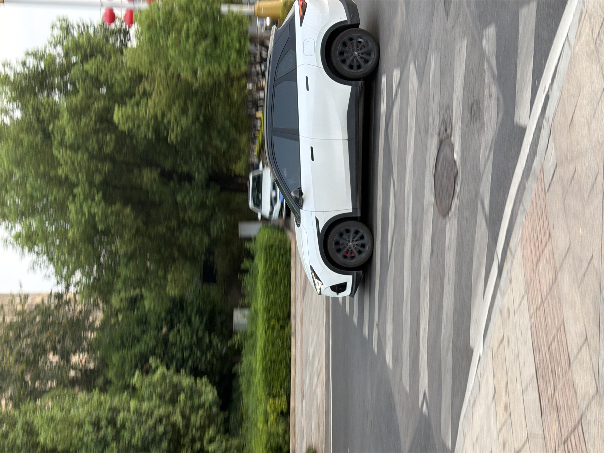 A white SUV with a black roof and trim is driving across a crosswalk in Jiangjin, China. The car is in motion, as indicated by the motion blur in the image. It is daytime, and the sun is casting shadows on the road. The background shows trees, buildings, and a lamppost with red lanterns. Another white car is parked in the background on the left. The crosswalk is clearly marked with white stripes on a gray asphalt road. A manhole cover is visible within the crosswalk. The sidewalk to the left of the crosswalk is paved with beige and pink tiles.