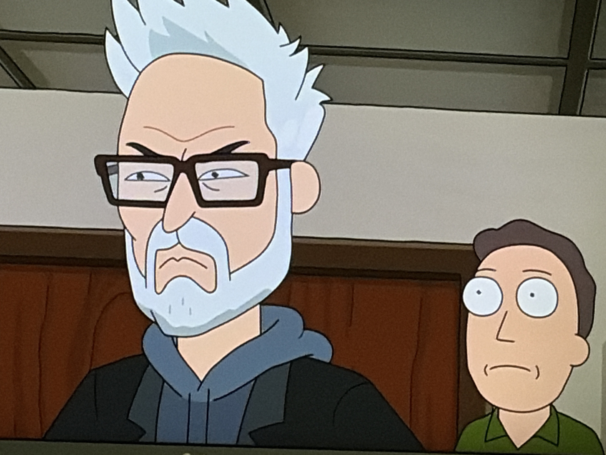 An animated scene features two male characters. The character in the foreground, on the left, is an elderly man with spiky white hair, a light blue beard, and light blue eyebrows. He is wearing thick-rimmed black glasses, a blue hooded sweatshirt under a dark jacket, and appears to have an annoyed or displeased expression. To his right and slightly behind him is a younger man with short brown hair and wide, circular white eyes. He is wearing a green shirt and has a neutral or slightly confused expression. They are both depicted from the chest up, against a background of what appears to be an interior setting with brown and beige walls. The image is a still from an animated series.