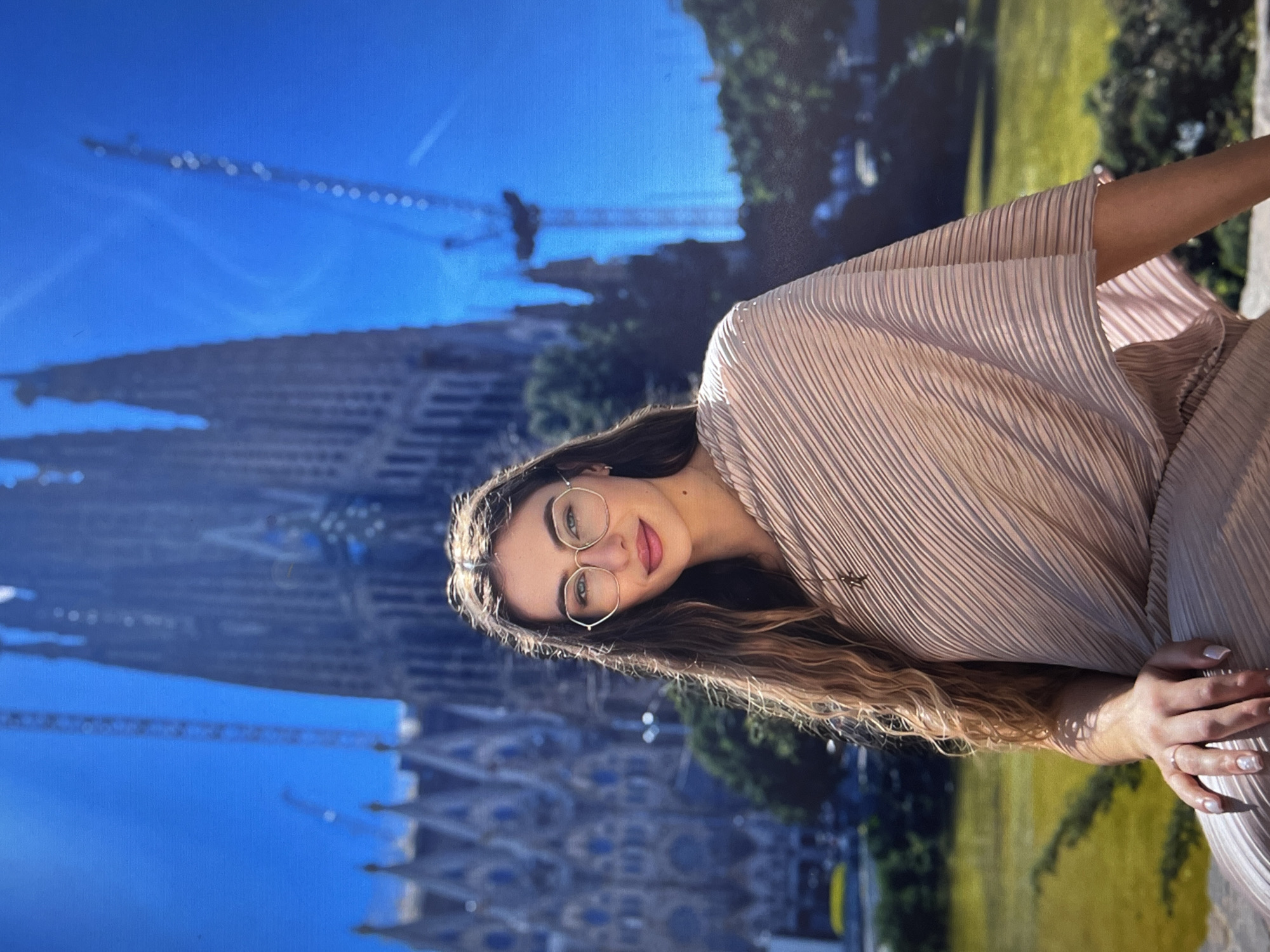 A woman wearing glasses and a light brown, textured outfit stands in front of a backdrop depicting the Sagrada Familia in Barcelona, Spain. The backdrop features a bright blue sky and construction cranes near the basilica. The woman's hair is long and wavy, and she is looking at the camera with a slight smile. Her hands are visible, adorned with light-colored nail polish.