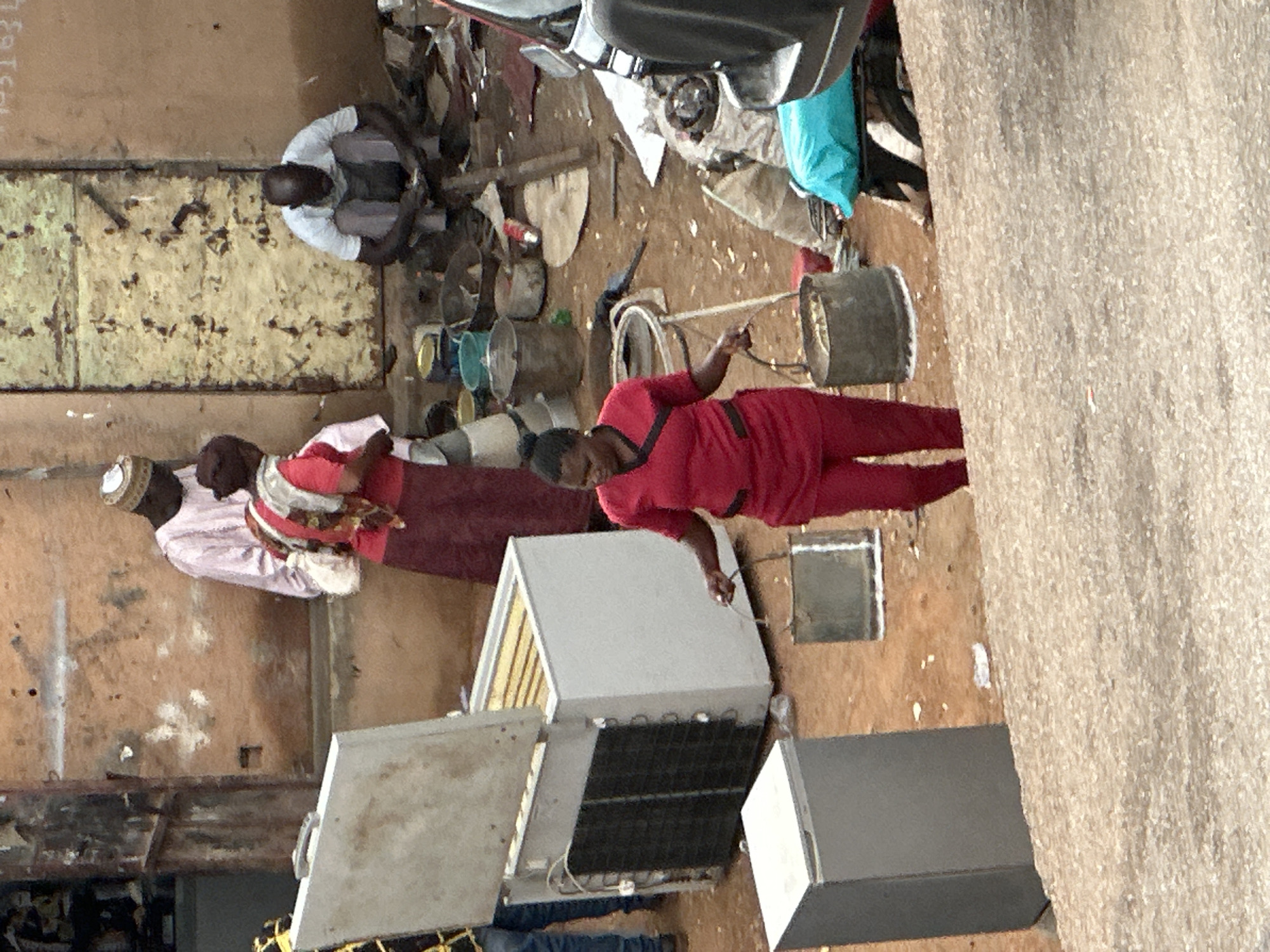 The image depicts an outdoor scene in Jos, Nigeria, likely a market or street-side area. In the foreground, a woman wearing a red uniform with black trim is carrying two metal buckets, one in each hand. She is walking on a gravelly incline. Behind her, a white refrigerator with its lid open stands next to another smaller, closed gray refrigerator. Further back, a man in a white shirt and grey trousers is squatting and appears to be handling something in his hands. To his left, another man in a light pink traditional outfit and a cap is standing. A woman in a red and white patterned scarf and a red dress is partially visible behind him. Various buckets, containers, and debris are scattered on the ground, suggesting a busy or possibly unorganized commercial area. A portion of a dark-colored vehicle is visible on the right side of the frame. The buildings in the background have rough, textured walls.