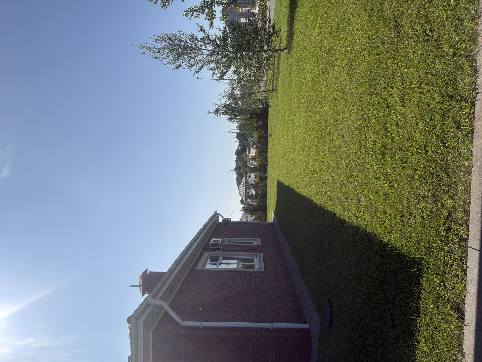 A brick house with a white gutter and downspout is visible on the left side of the image. The house has two windows and an air conditioning unit mounted on the exterior wall. A chimney is located on the roof. The house casts a long shadow across a well-maintained green lawn. Several trees of varying sizes are scattered across the lawn, with streetlights visible in the background. The sky is clear and blue, with the sun shining brightly. The scene appears to be in the city of Laskino, Russia.