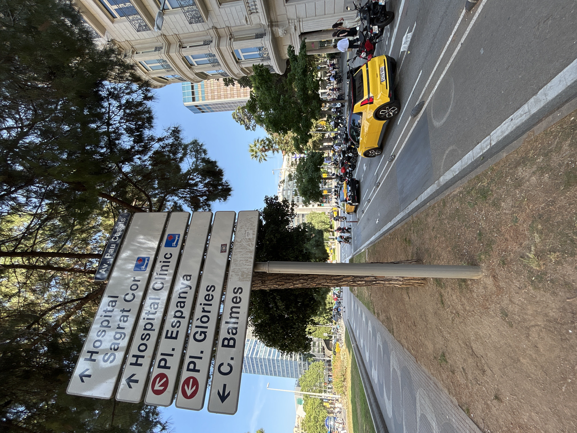 The image depicts a street scene in Barcelona, Spain. The camera is positioned at street level, angled upwards, and rotated counterclockwise.
In the foreground, there's a signpost with multiple directional signs pointing towards different locations. The signs read: "Hospital Sagrat Cor," "Hospital Clínic," "Pl. Espanya," "Pl. Glòries," and "C. Balmes," each with an arrow indicating the direction. The signpost is next to a sidewalk.
Beyond the signpost, a street stretches into the distance. Several vehicles, including a yellow car and numerous motorcycles, are parked along the street. People are visible near the vehicles.
Buildings are on the right side of the street. Trees line the street. The sky is blue.