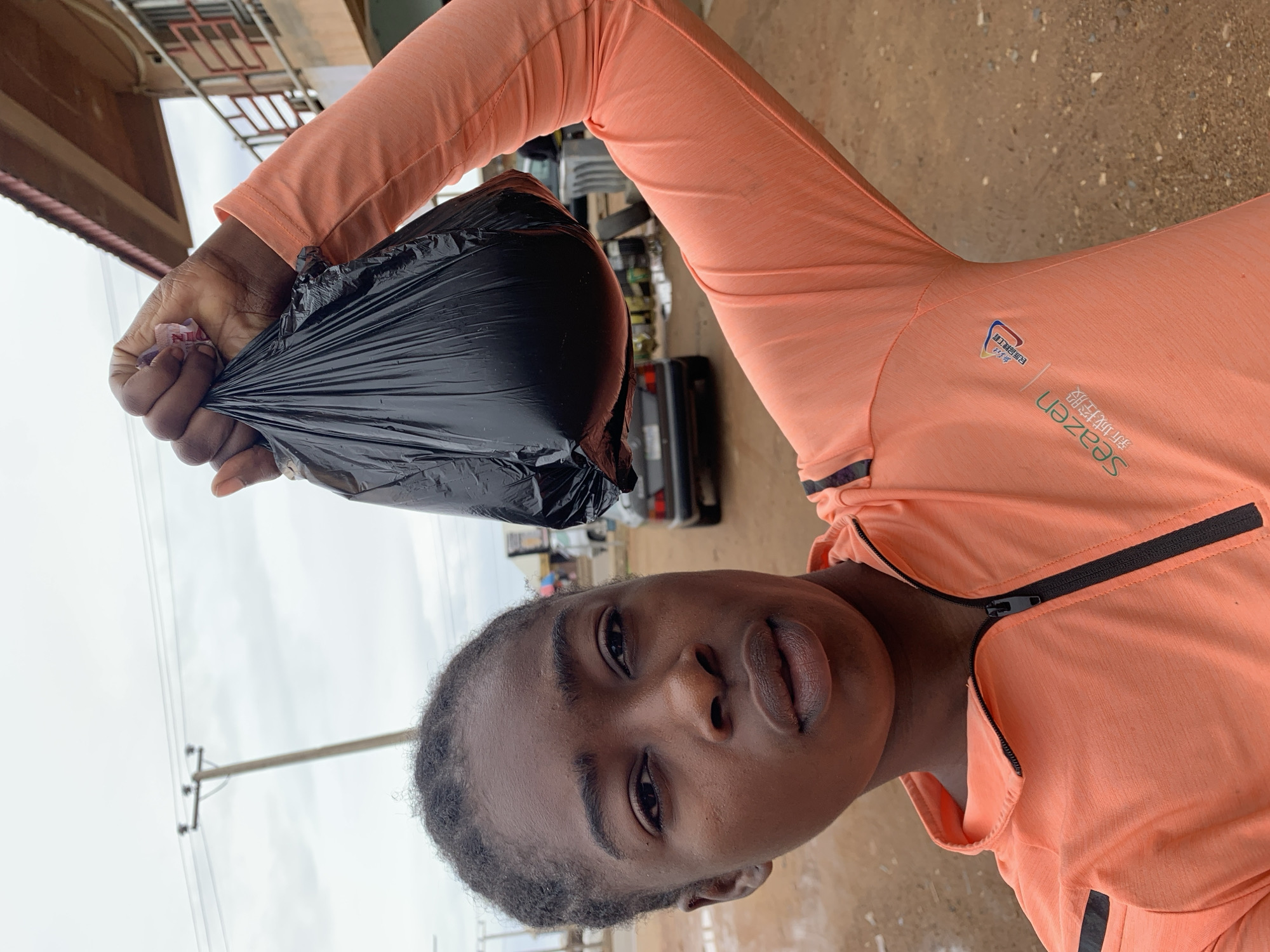A person with dark skin and hair stands outside in Rafin Par, Nigeria. The person holds a black plastic bag with their right hand. They are wearing an orange long-sleeved athletic shirt with a black zipper and the words "Seazen" and some other writing in a different language. Behind the person are buildings, power lines, a car, and other objects typical of a city street. The ground is a light brown color. The sky is overcast.