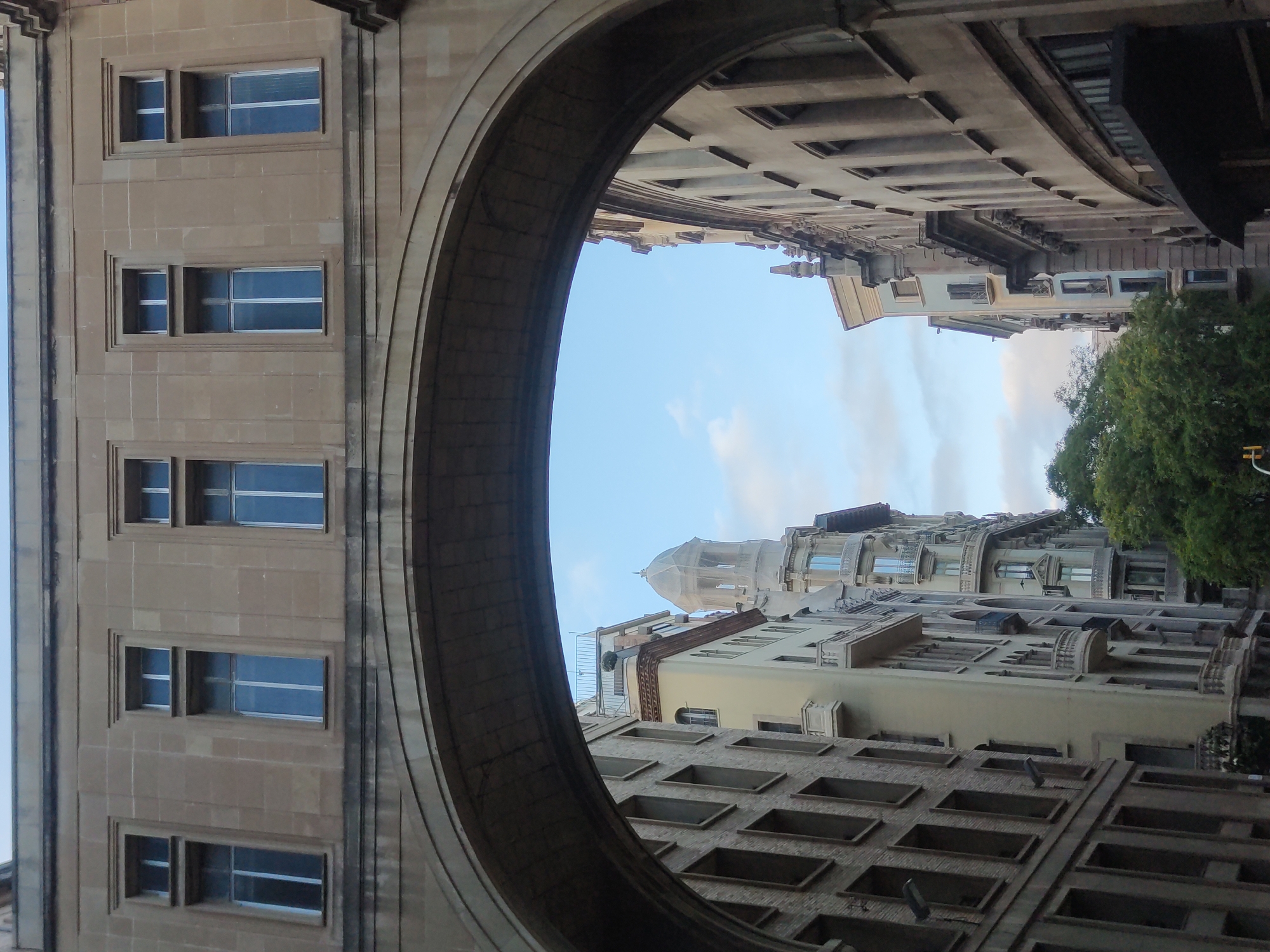 The image captures an urban street scene in Barcelona, Spain, viewed from beneath a large architectural archway. The arch frames a view of several historic buildings lining a street. The buildings exhibit a variety of architectural styles, with some featuring ornate facades, balconies, and arched windows, while others appear more utilitarian with rows of rectangular windows. A prominent domed structure, possibly a church or a historic public building, is visible in the distance. The sky is partly cloudy, indicating daytime. There are no people visible in the frame, nor any specific actions or interactions. The focus is on the architecture and the urban environment.