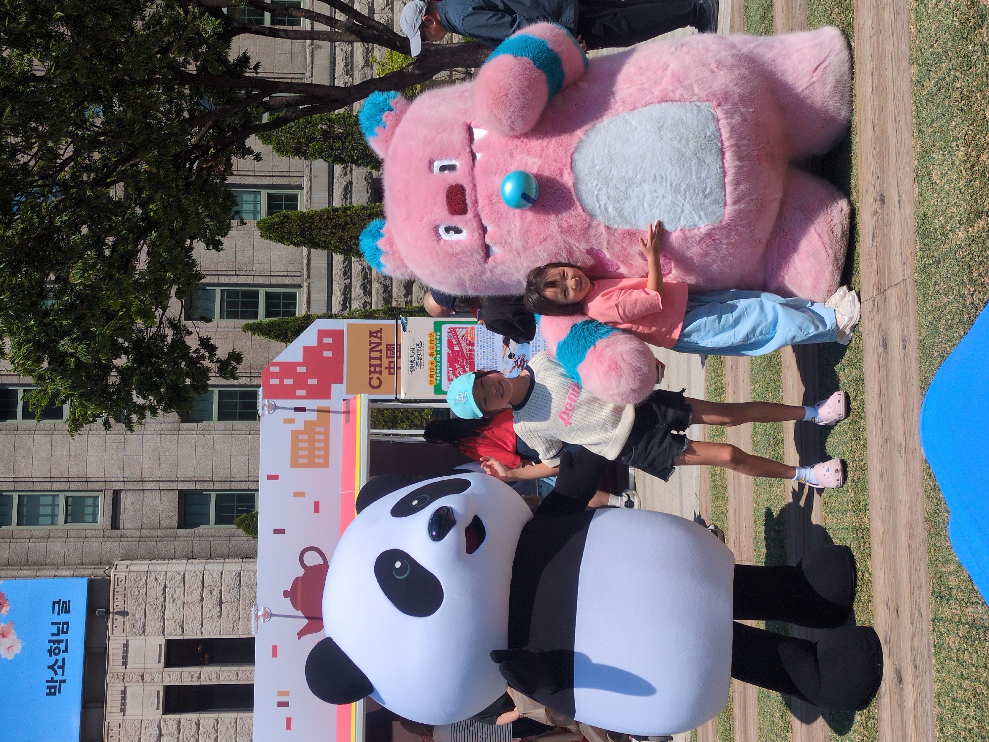 Captured in an outdoor setting, possibly a park or event space within Seoul, South Korea, the image features two individuals posing with oversized mascot figures. A girl in a peach shirt and light blue pants stands next to a large, pink, plush-like creature with blue accents, possibly a mascot for an event. Another child, sporting a light-colored shirt, dark shorts, and a blue cap, is posing with a panda mascot. Behind them, there is a backdrop labeled "CHINA" in large, bold letters, with Chinese characters and images related to traditional arts and crafts. To the left, a banner in Korean is visible, further indicating the location. The background also includes a stone building with windows, suggesting the location may be near a government building or institution. The lighting suggests it is daytime with fairly clear skies. People are standing in the background, showing an organized event.