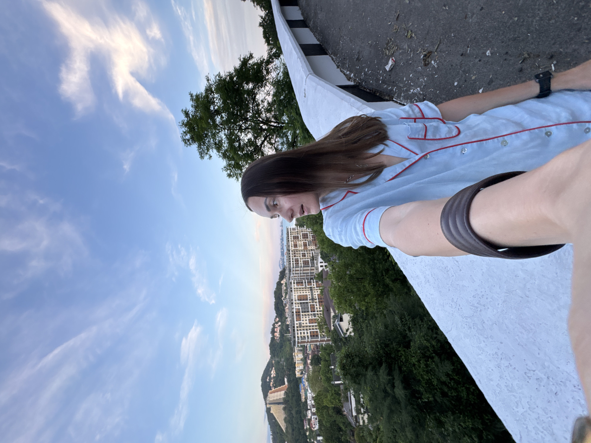 A woman with long brown hair is taking a selfie while standing on a paved area next to a white barrier wall. She is wearing a light blue pajama top with red trim. Behind her, a city sprawls across a hillside, leading down to a body of water. Buildings of various sizes and architectural styles are visible, interspersed with dense green trees. In the background, hills rise up, topped with structures. The sky is a mix of blue with wispy white clouds. A large green tree stands to the right of the woman. The ground beside the barrier wall is a paved road with scattered debris.
