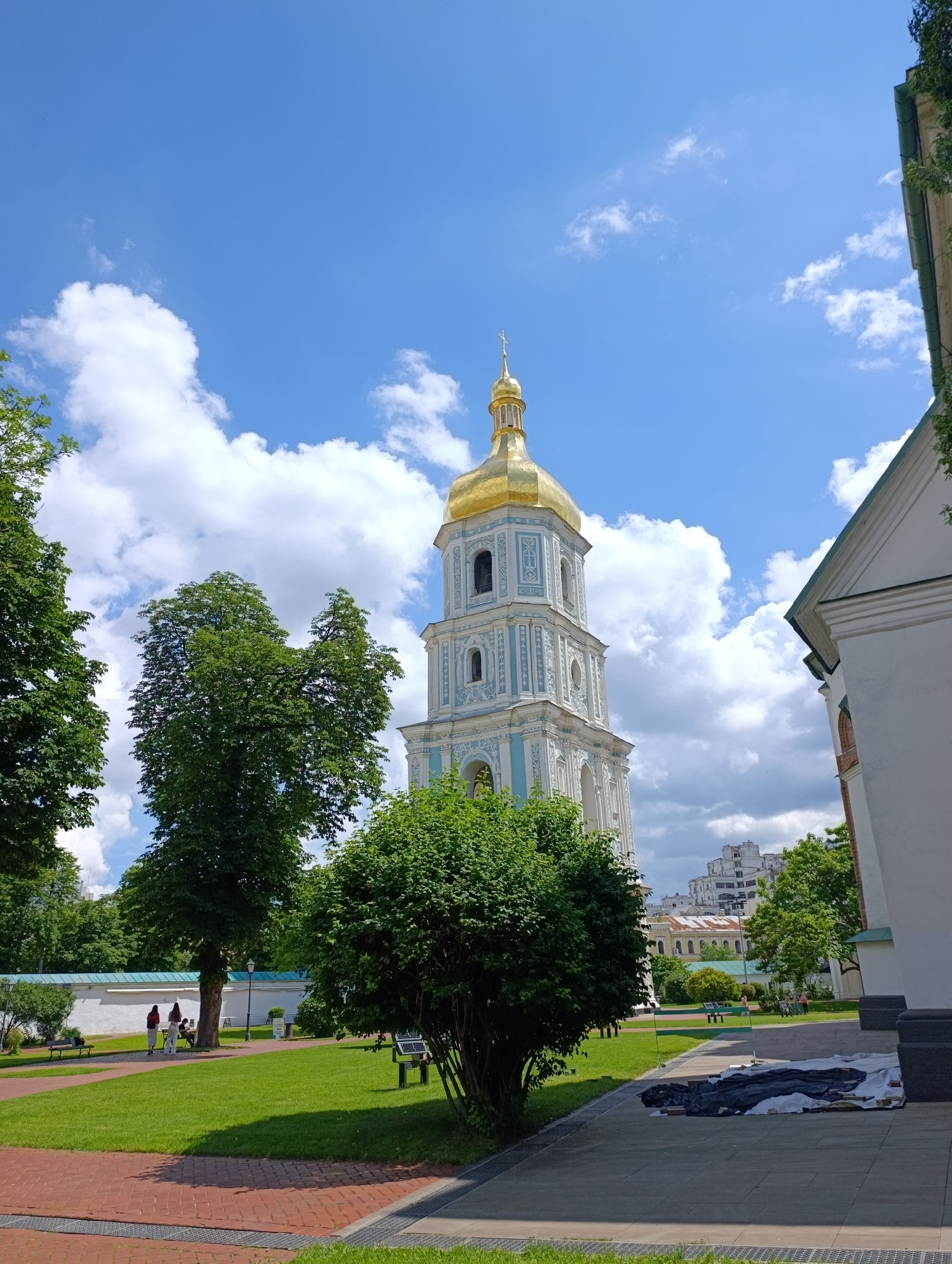 The image shows a bell tower of Saint Sophia Cathedral in Kyiv, Ukraine, on a sunny day with a blue sky and white clouds. The bell tower is white with blue accents and a golden dome topped with a cross. In the foreground, there is a green lawn with trees and a paved walkway. In the background, there are other buildings and more trees. Several people are visible walking on the lawn and sitting on benches.
