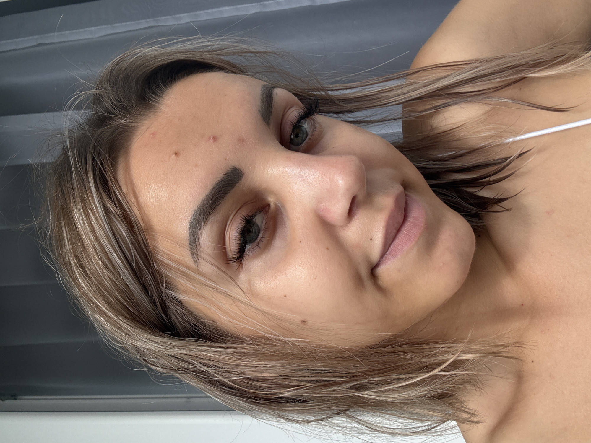 The image is a close-up, head-and-shoulders portrait of a young woman with medium-length blonde hair, some of which appears wet. She is looking slightly off-camera to her right with a subtle smile. Her skin is light-toned, and there are a few blemishes visible on her forehead and cheek. She is wearing a white strap, possibly from a top or bra, visible across her chest. The background consists of a dark gray curtain and a lighter gray vertical element, possibly a door frame or wall. The lighting is soft and natural, coming from the left side of the frame. The scene appears to be indoors. There is no information about the city of Kholmsk, Russia, or any other specific location.