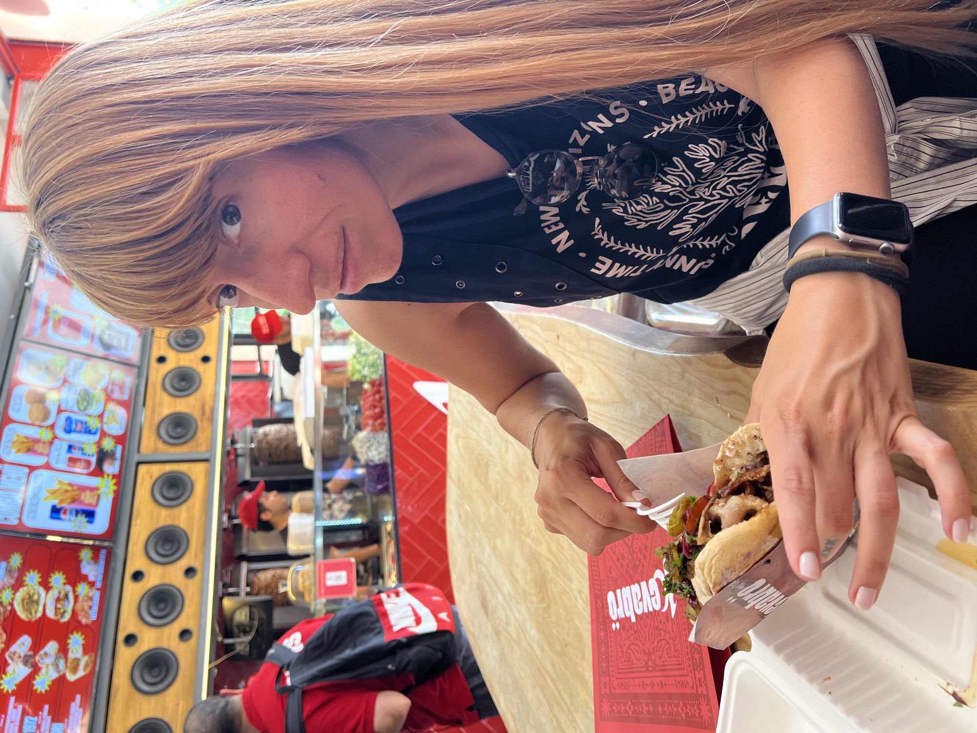 An outdoor shot captures a woman sitting at a wooden table, holding a food item, and looking to the side with a slight smile. She has long, light brown hair with bangs, and she is wearing a black tank top with silver grommets and a graphic print. Sunglasses are hanging from her neck. A smartwatch is on her left wrist. The background shows a food establishment with a menu board displaying various food items, along with speakers mounted above a counter where food is being prepared. A person in a red shirt and backpack is visible to the left, and another person in a red cap is working behind the counter. The overall scene suggests a casual dining experience in a lively urban setting.