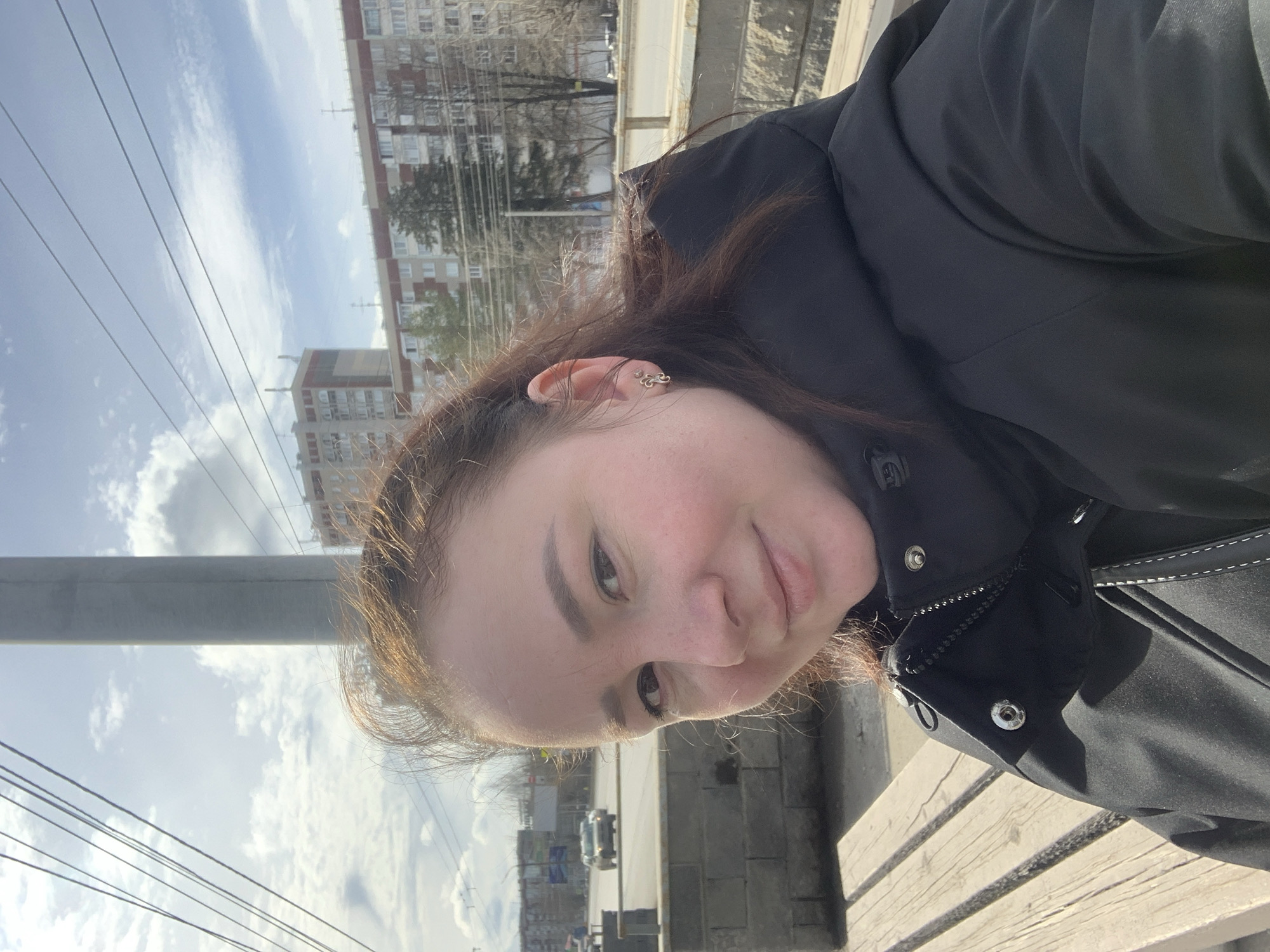A person is facing the camera, positioned outside in an urban setting. The person is wearing a black jacket. Buildings can be seen in the background, along with power lines. A street with vehicular traffic is also visible. Based on the architecture and available information, the location is potentially within the city of Izhevsk, Russia.