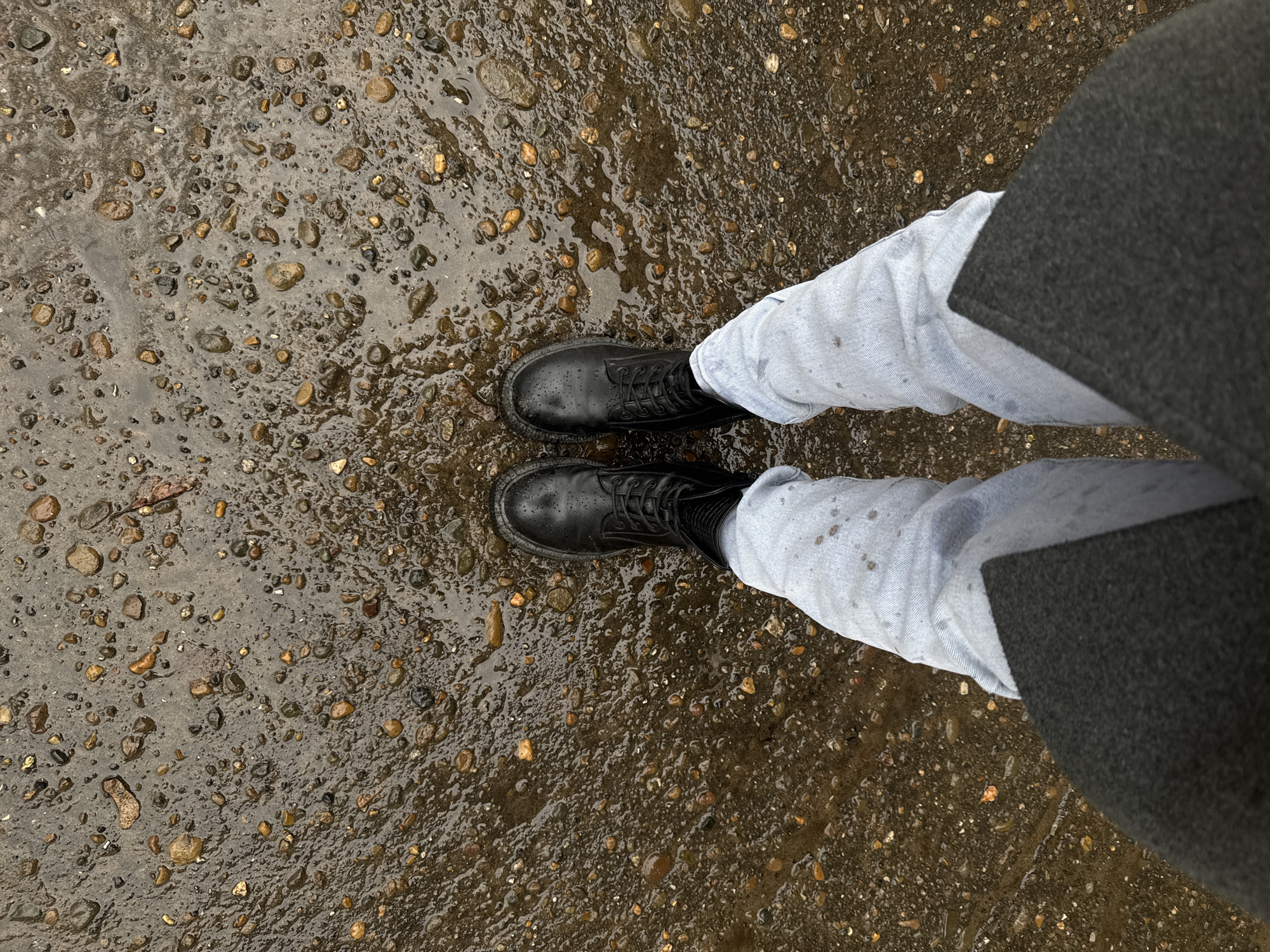 The image presents a first-person perspective of a person standing on a wet, gravelly surface. The person is wearing black, lace-up boots, light blue jeans, and a dark gray coat or jacket. The ground is covered with small pebbles and is glistening with water, indicating it has recently rained or is still raining. The location is unknown. There are no other people or subjects visible in the image. The person is simply standing still, presumably taking the picture. There is no visible interaction with the environment other than the boots making contact with the ground. There are no indications of Chaikovsky, Russia.