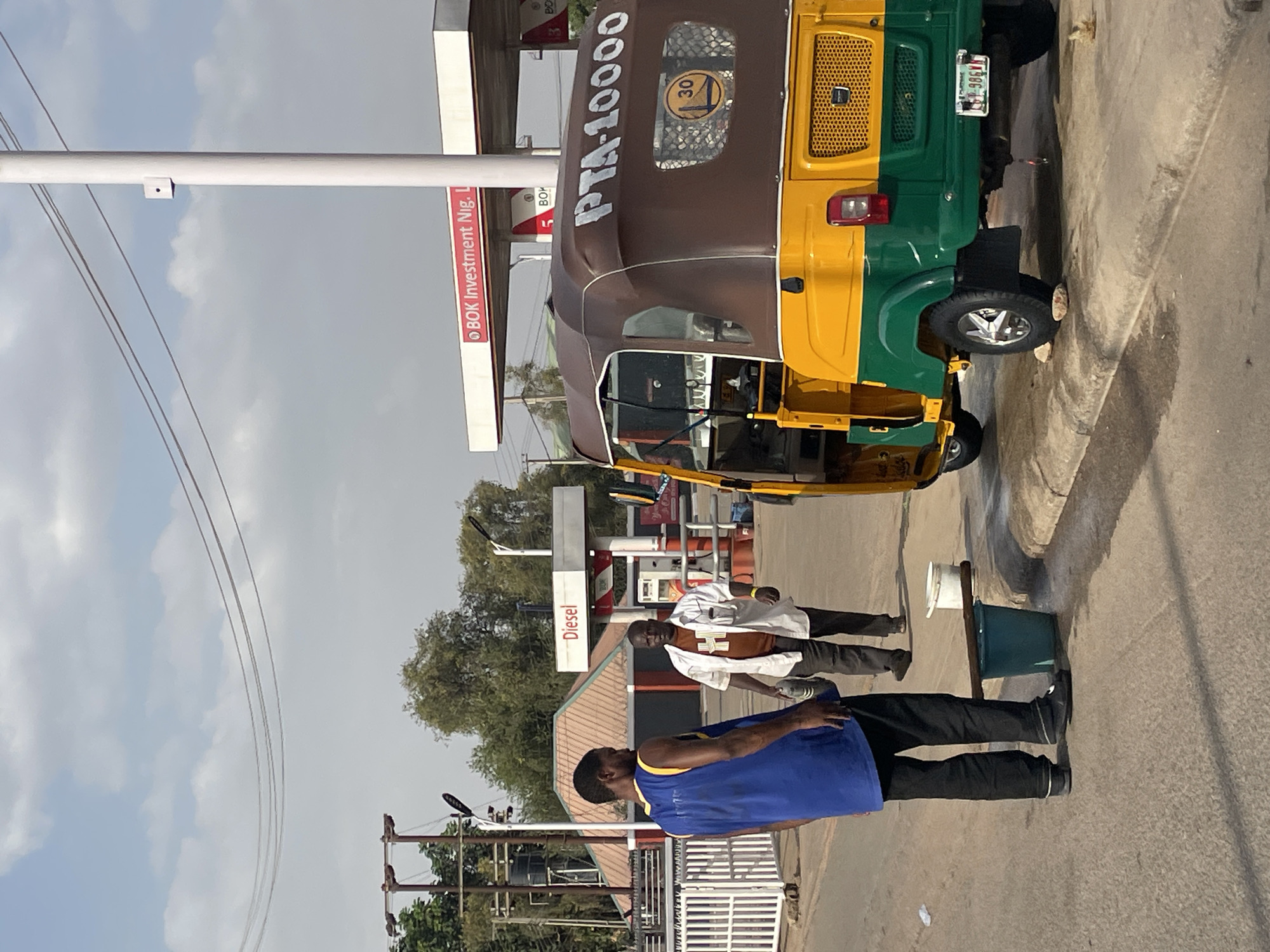 The image depicts a street scene in Jos, Nigeria, featuring a "keke" (tricycle taxi) with the markings "PTA-10000" and a 30 km/h speed limit sign. The keke is colored brown, yellow, and green. A man wearing a blue shirt and black pants is standing near a bucket. Another man in a white shirt and dark pants is walking in the background. The setting includes a petrol station with visible "Diesel" signage and "BOK Investment Nig. Ltd" branding. Utility poles with electrical wires are visible, with trees and a cloudy sky in the background. The scene appears to be along a paved road.