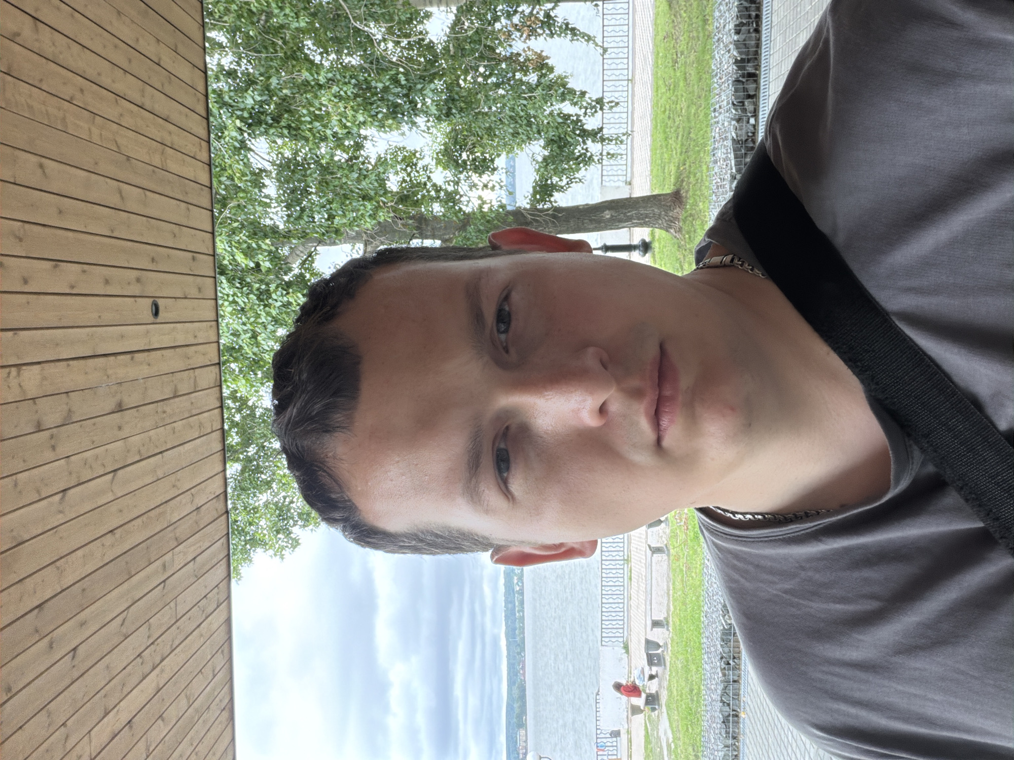 A selfie is taken by a young man with short, dark hair and a serious expression, facing the camera directly. He is wearing a gray t-shirt and a silver chain necklace. A black strap from a bag is visible across his shoulder. He is standing outdoors, under a wooden canopy. Behind him, a body of water, possibly a river or lake, is visible under a cloudy sky. Trees with green leaves line the background, and a paved walkway with benches and grassy areas is present. In the distance, a person in a red shirt is seated on a bench near the water. The setting appears to be a park or embankment within the city of Votkinsk, Russia, given the context of the image originating from Votkinsk.